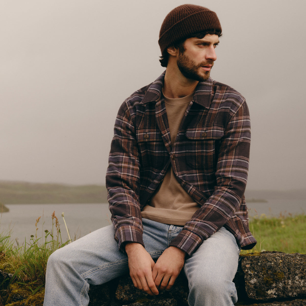 A man wearing a brown knit beanie, a brown and black plaid flannel shirt over a beige t-shirt, and light blue jeans, sitting outdoors on a stone wall with a misty lake in the background.