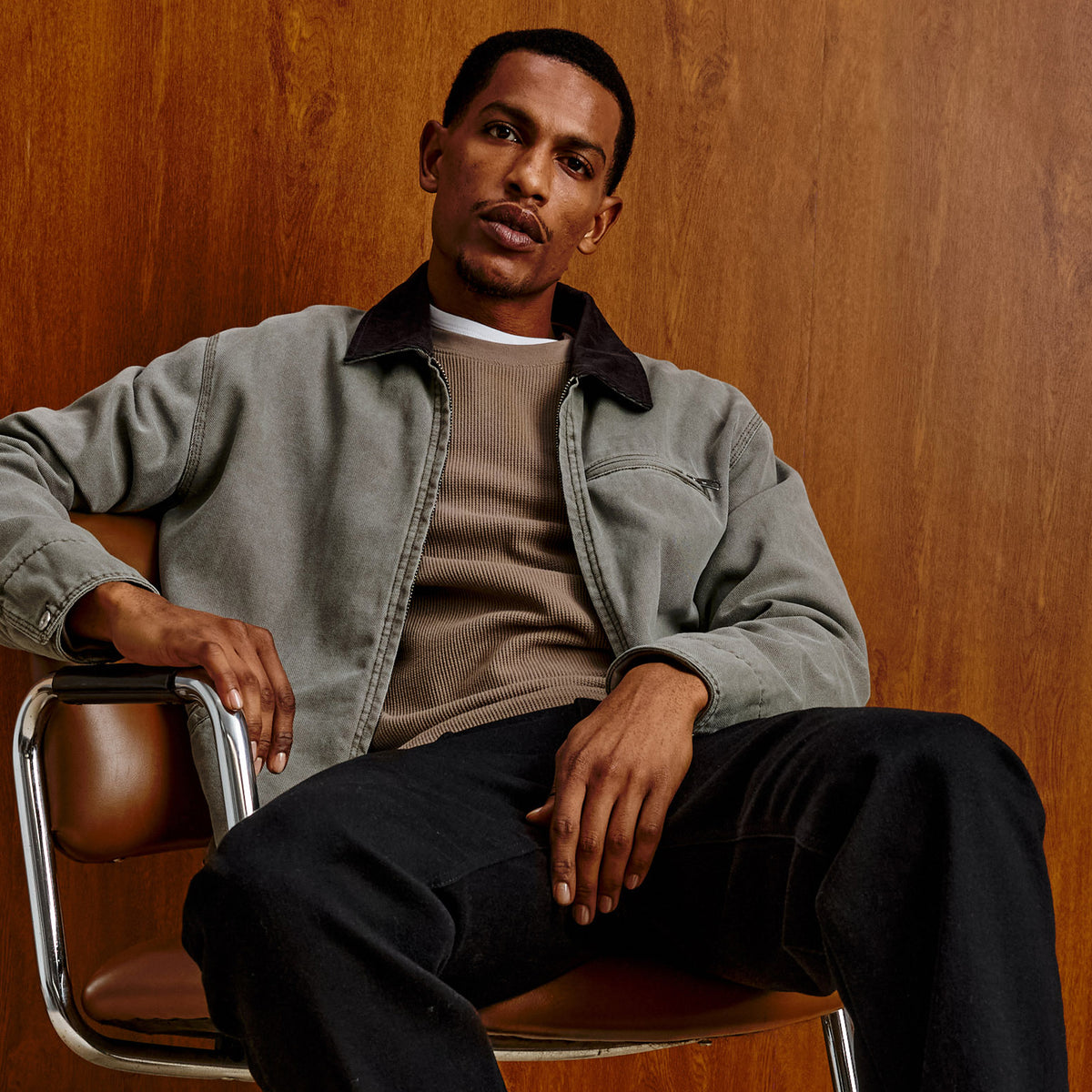A man wearing a muted gray-green overshirt over a taupe knit sweater and black pants, seated on a brown leather chair against a warm wood-paneled wall.