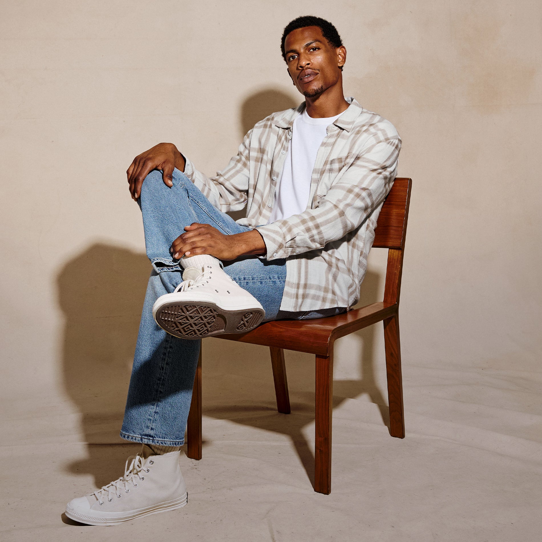 A man sits on a wooden chair wearing a beige and white plaid button-up shirt over a white t-shirt, blue jeans, and white high-top sneakers against a warm beige studio backdrop.