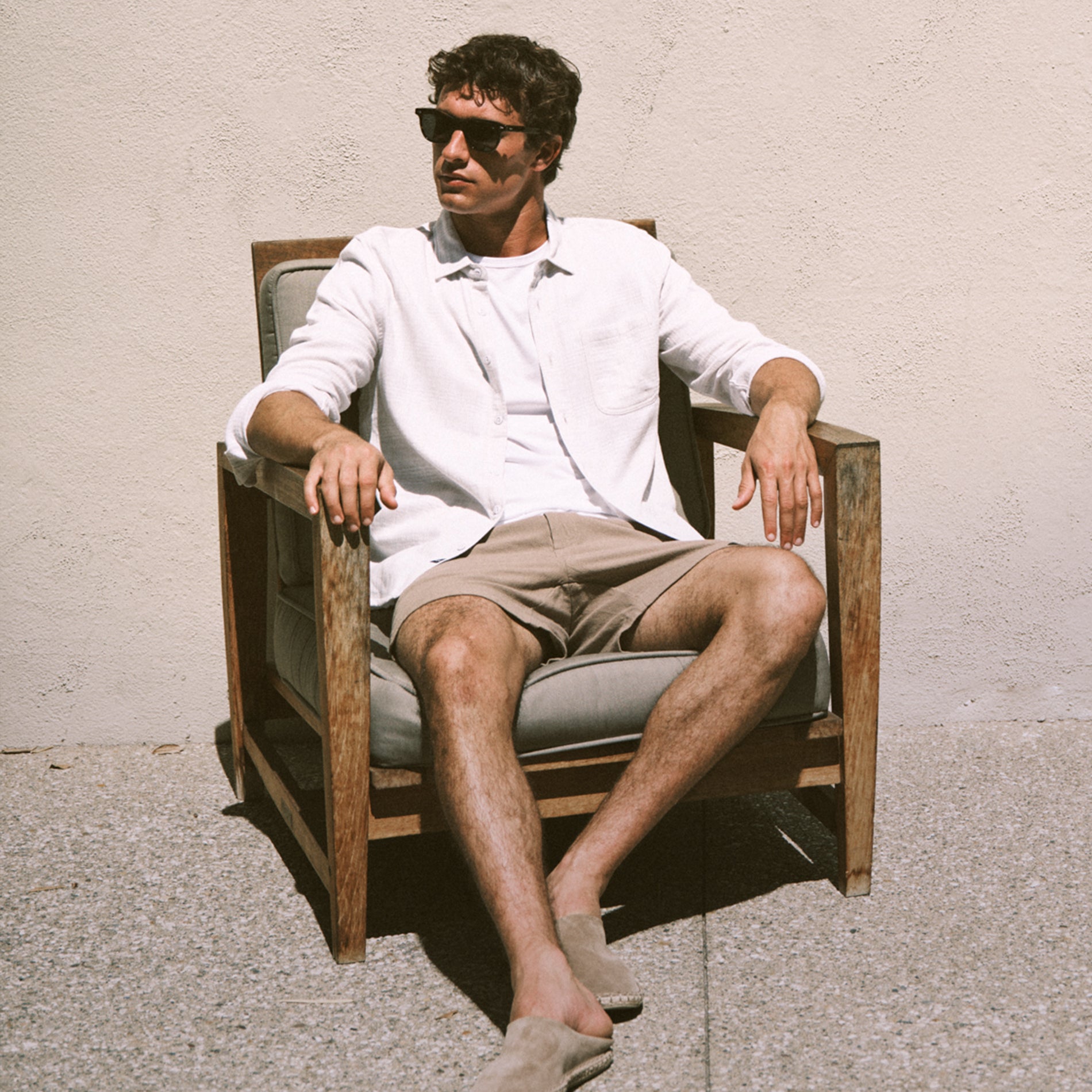 Man sitting on a chair wearing sunglasses and casual clothing against a plain wall.