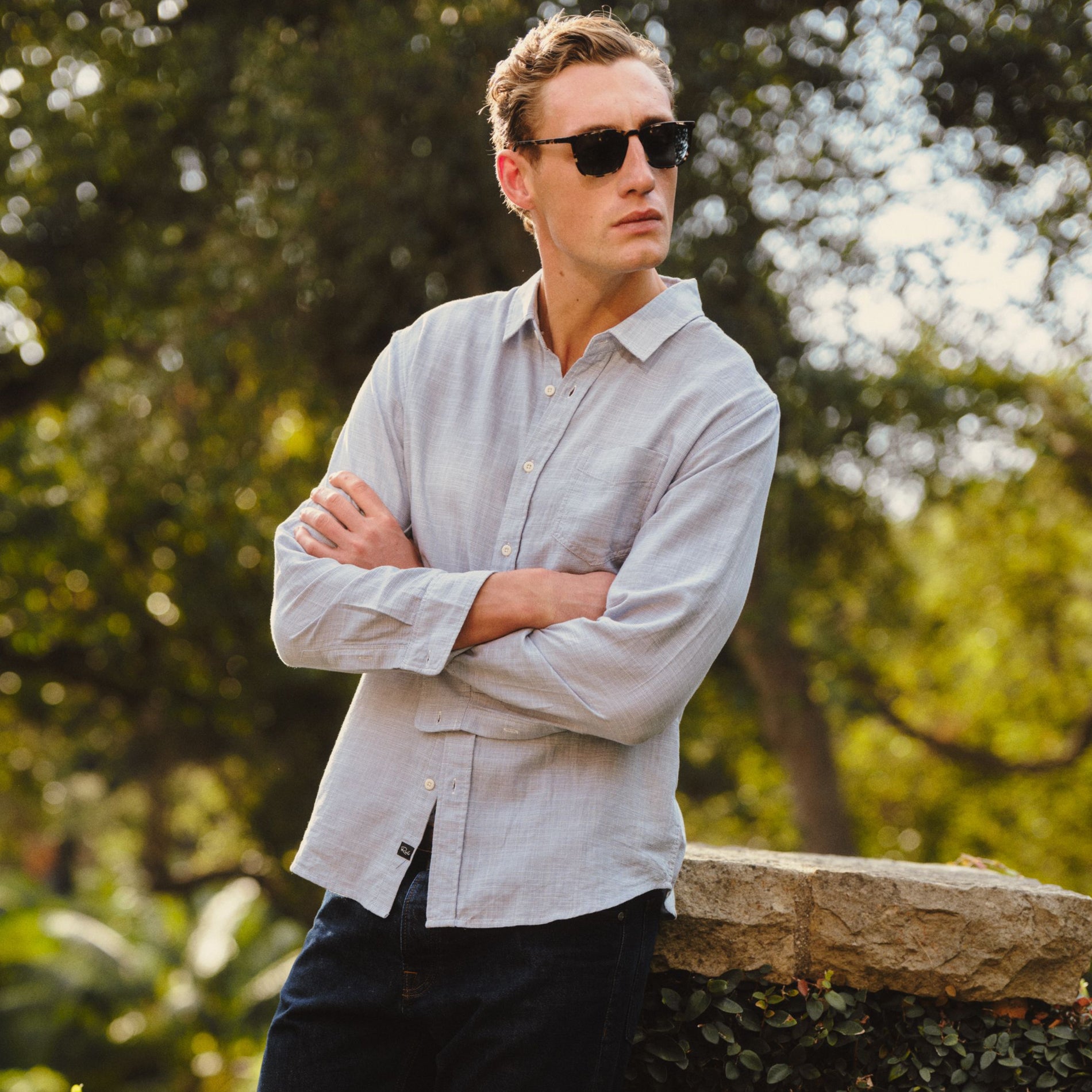 A man wearing a light gray long-sleeve linen button-up shirt and dark sunglasses, standing outdoors with arms crossed in front of a stone wall and green trees.
