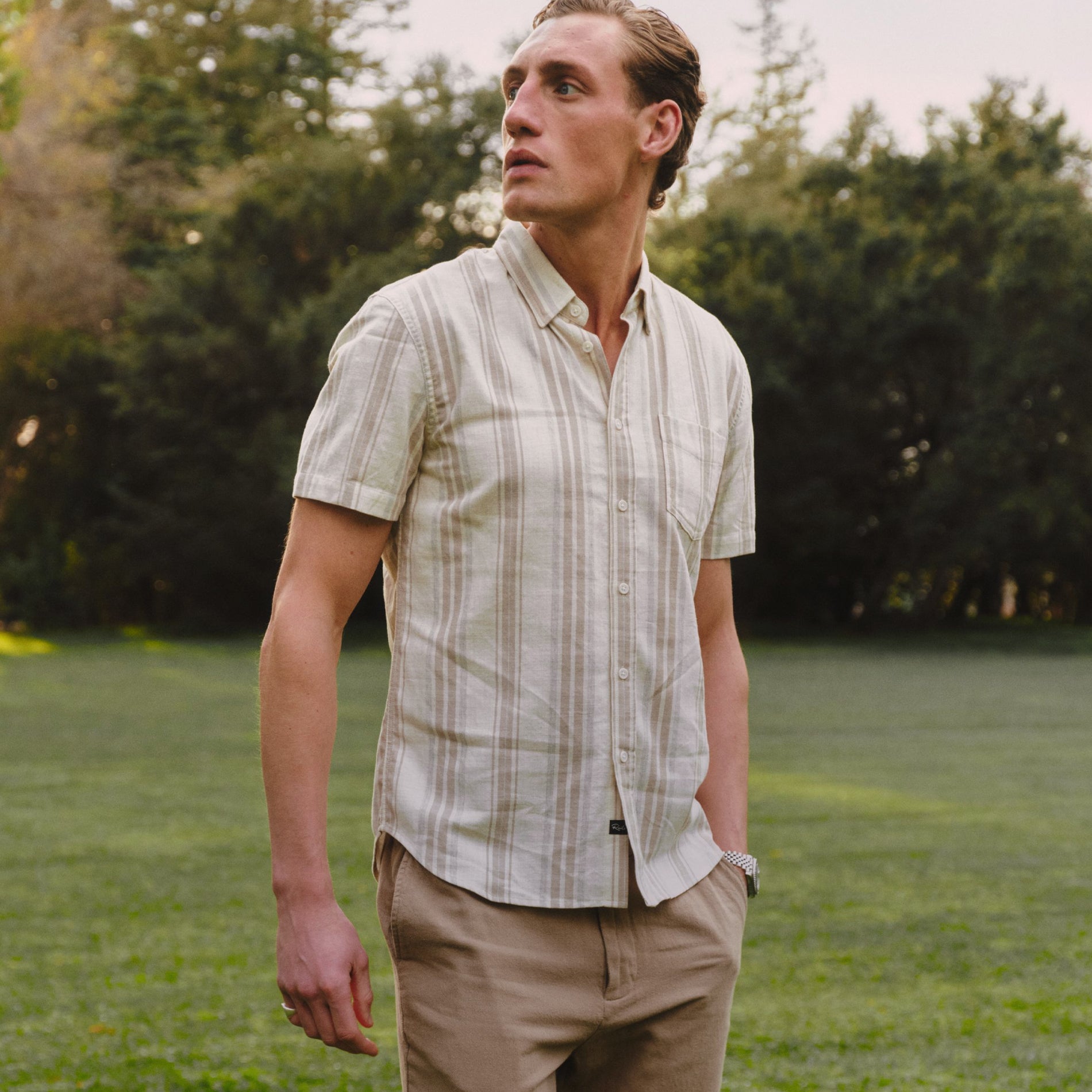 A man standing outdoors on a grassy lawn wearing a short-sleeve button-up shirt with vertical beige and white stripes, paired with khaki pants.