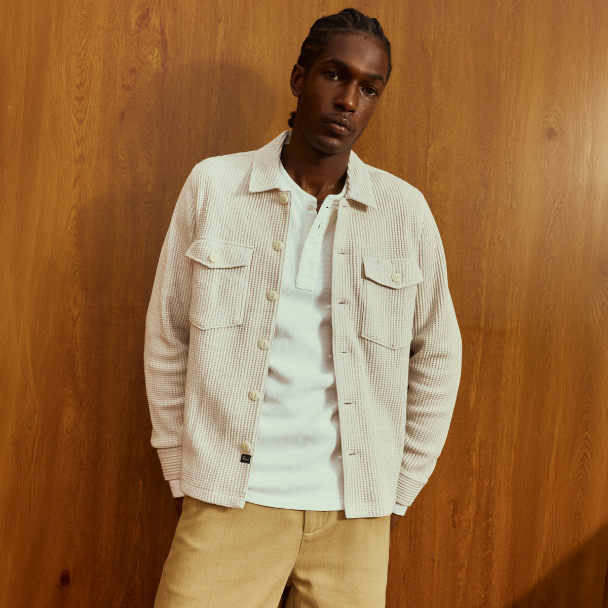 A man wearing a light beige textured overshirt over a white polo shirt and tan pants, standing against a warm wood-paneled wall.