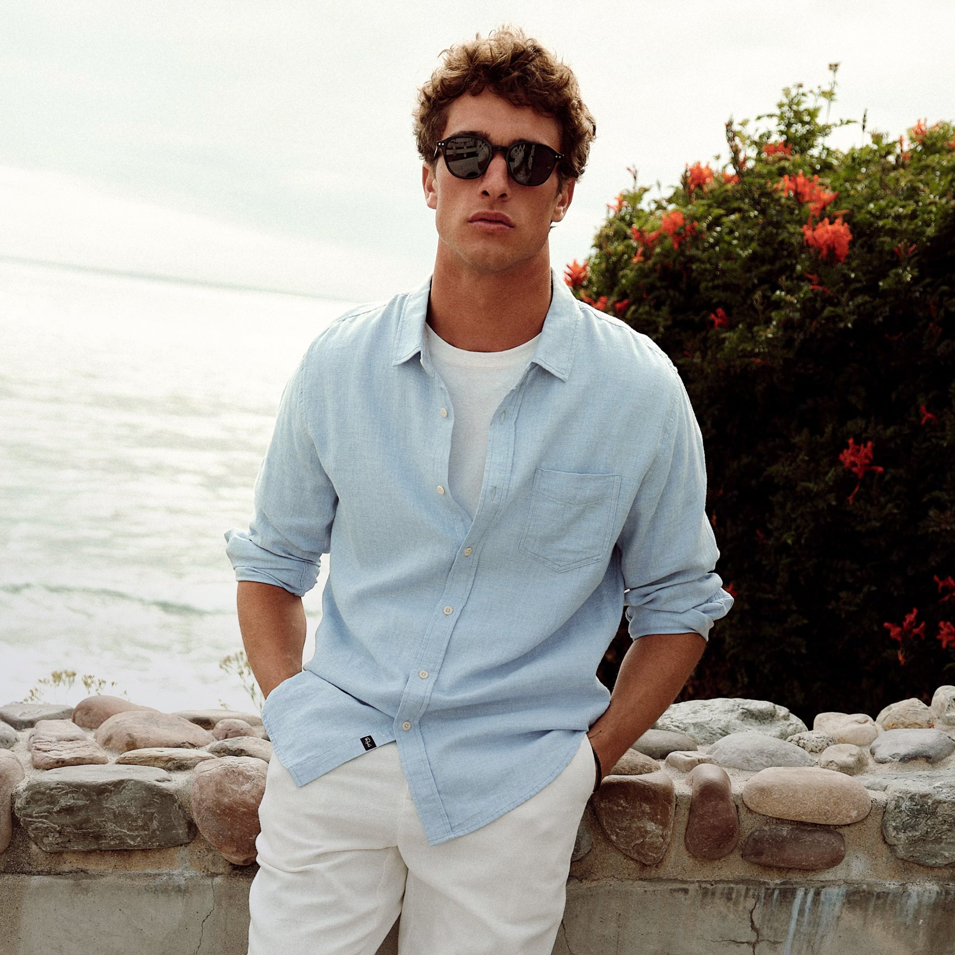 Man wearing sunglasses and a light blue shirt by a body of water with greenery.