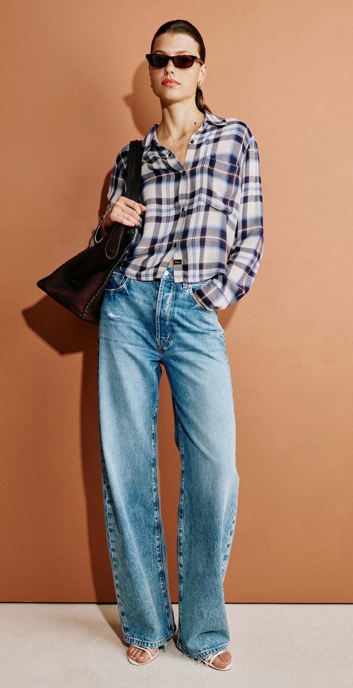 A model wearing a blue and white plaid flannel shirt, light-wash wide-leg blue jeans, and carrying a black shoulder bag, shown in a full-body shot and a close-up panel against a warm terracotta background.