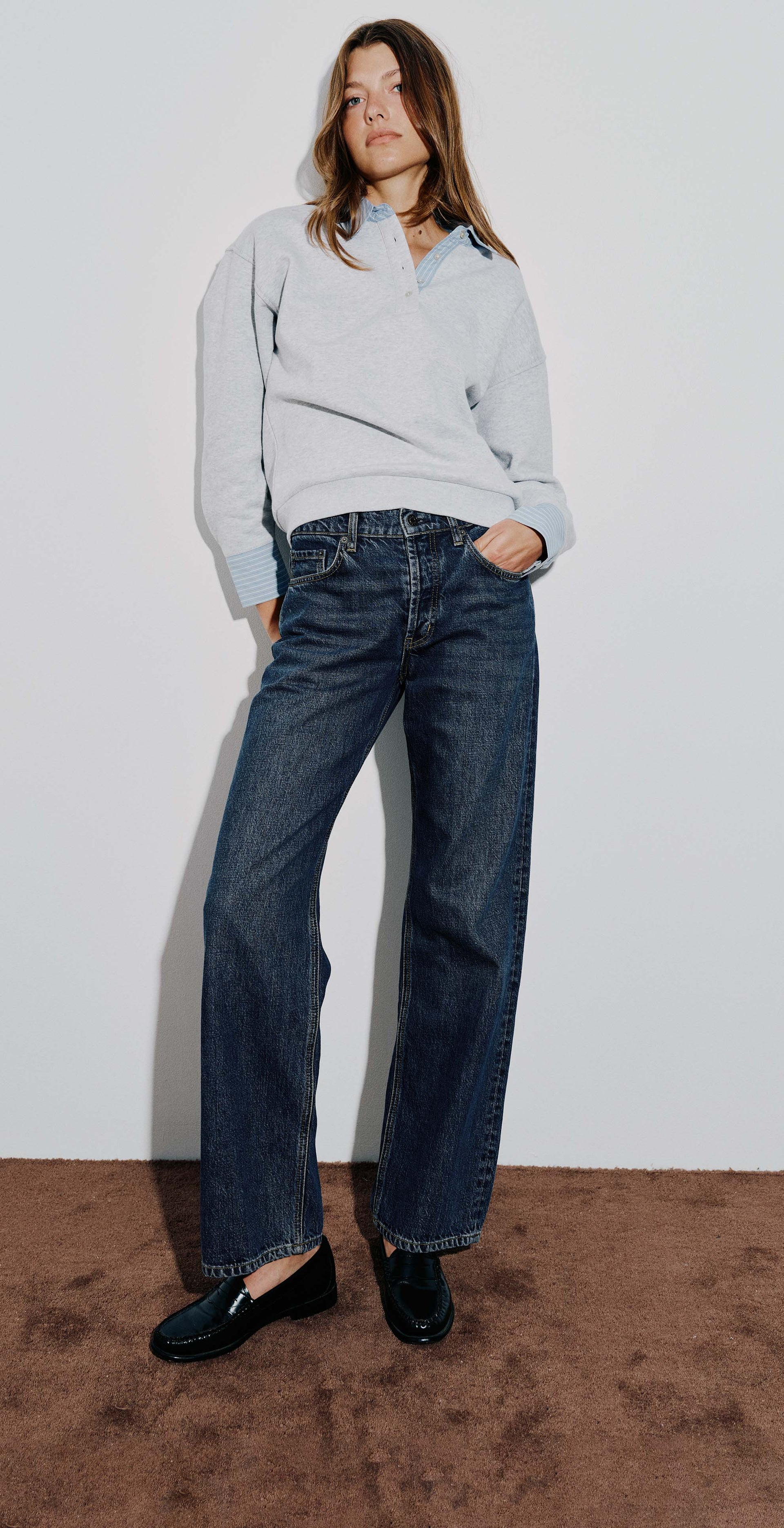 Three-panel lifestyle photo of a woman modeling dark blue wide-leg jeans with a light gray long-sleeve shirt and black loafers, posed against a white wall with a brown carpeted floor.