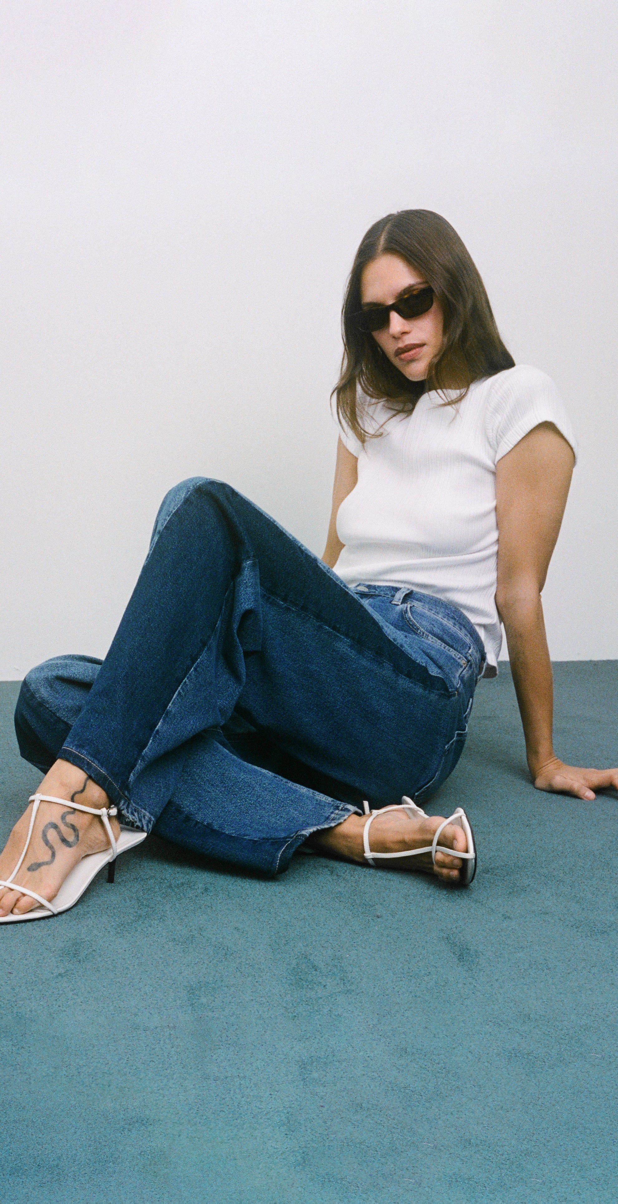 Woman sitting on a blue carpet wearing sunglasses, white shirt, blue jeans, and white heels.