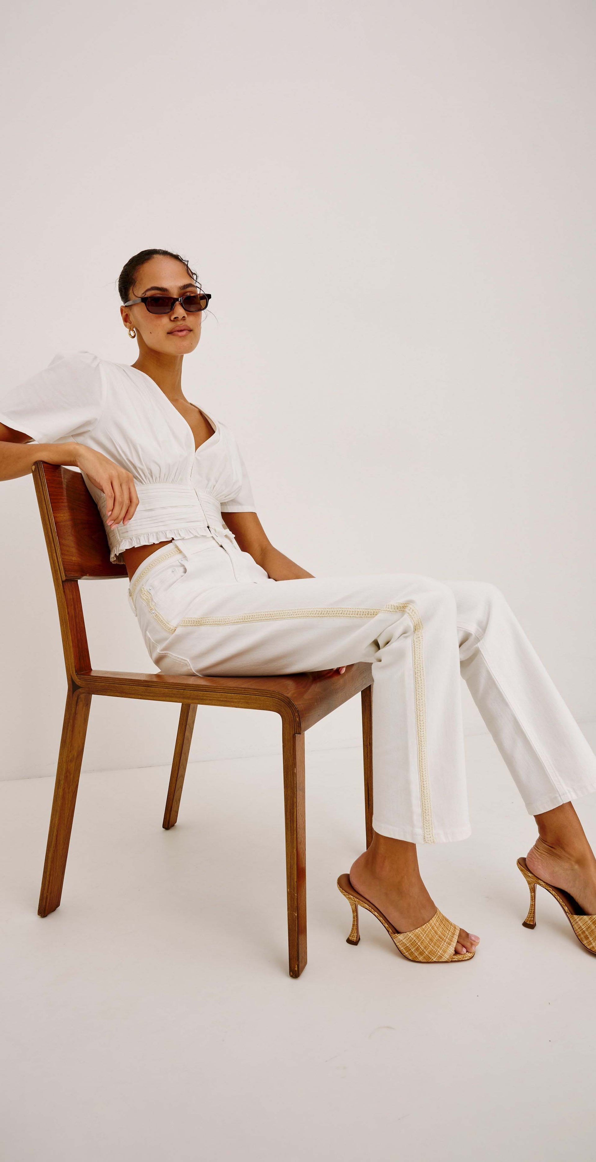 A woman wearing a white short-sleeve top and white wide-leg trousers sits on a wooden chair against a plain light gray studio background. She is wearing gold heeled sandals and dark sunglasses.