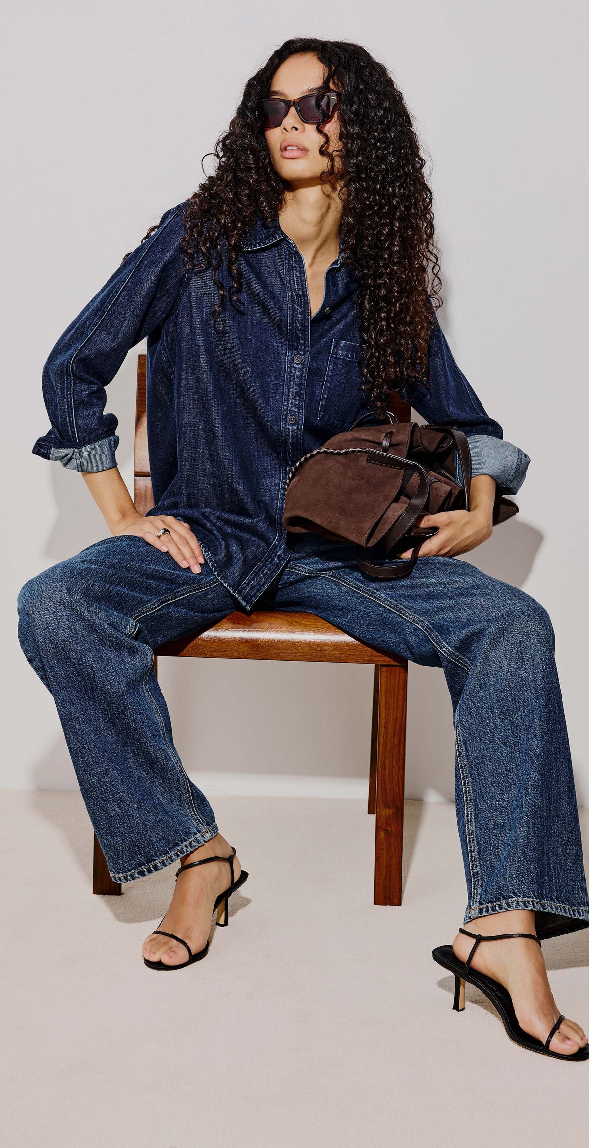 A split-screen fashion photo showing a woman modeling a dark blue denim shirt and wide-leg blue jeans in two poses against a plain light gray studio background. She is holding a brown shoulder bag and wearing black heeled sandals.