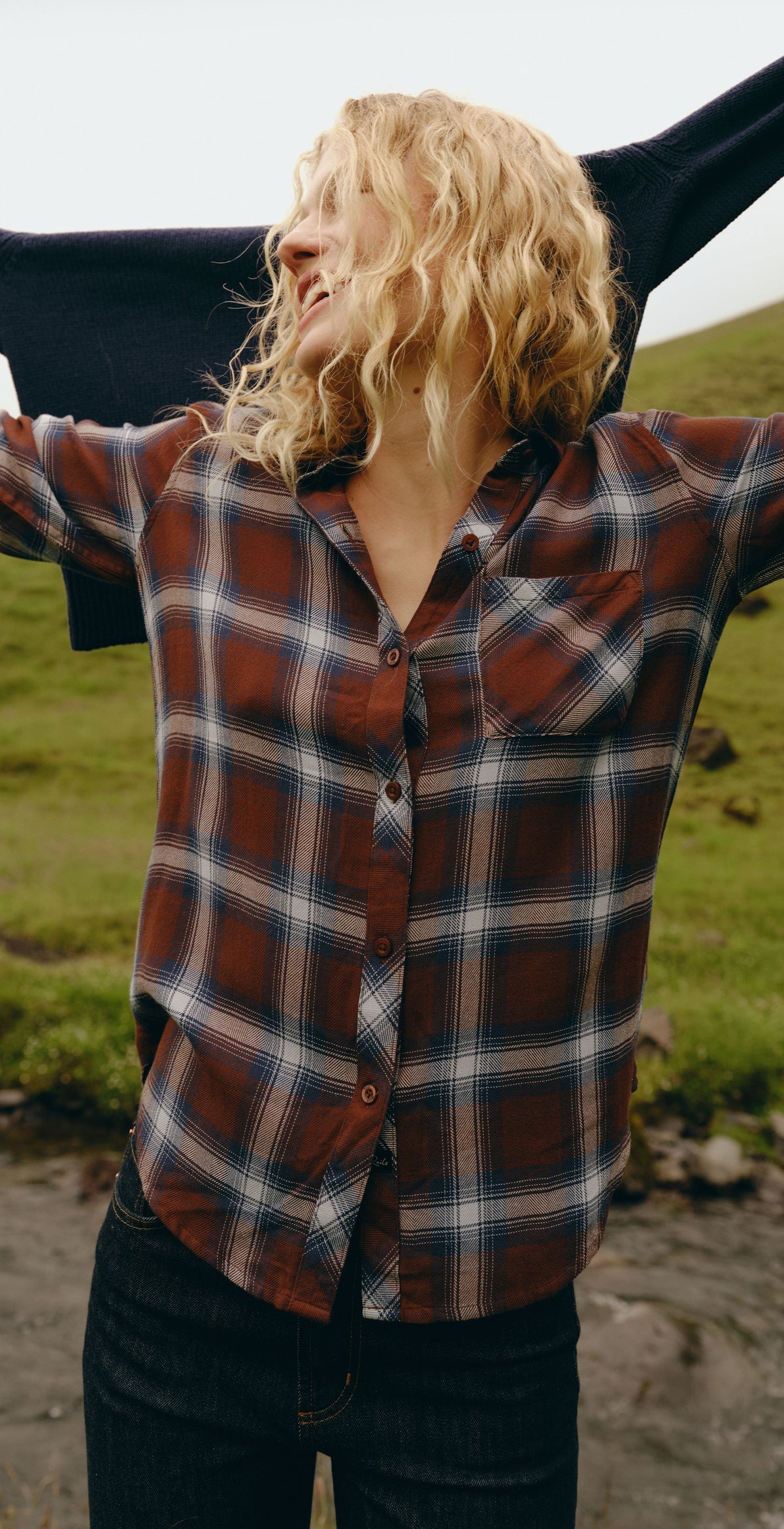 A three-panel lifestyle photo of a woman outdoors wearing a red, black, and white plaid button-up shirt, paired with a dark blazer and jeans, standing on a grassy hill.