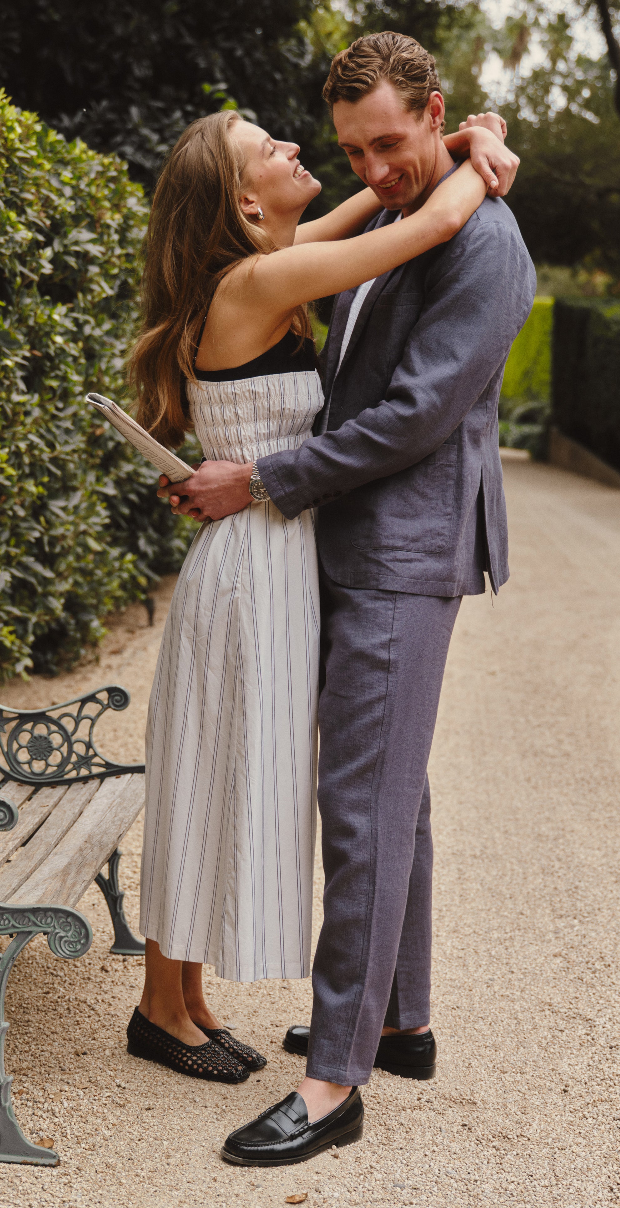 Woman in a white dress posing outdoors with a bench and greenery.