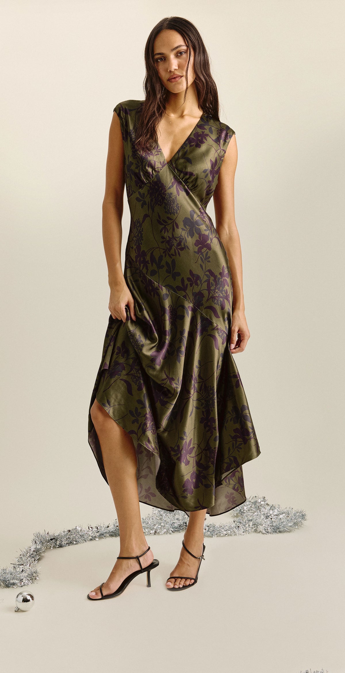 A woman modeling a sleeveless dark olive green midi dress with a subtle floral pattern, shown in two studio poses against a light beige background with decorative silver tinsel.