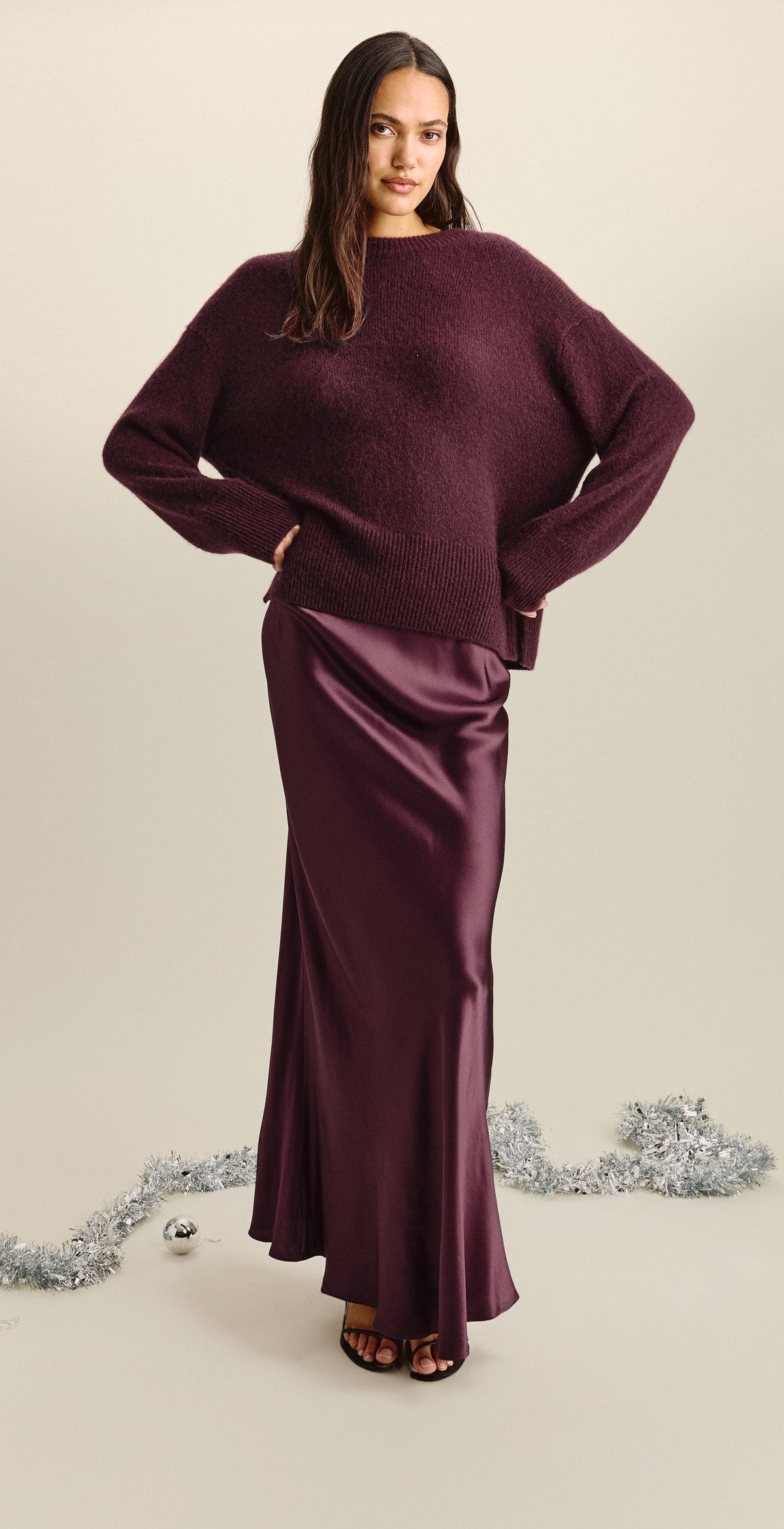 A woman modeling a matching burgundy knit sweater and satin maxi skirt set against a light beige studio background with silver tinsel on the floor.