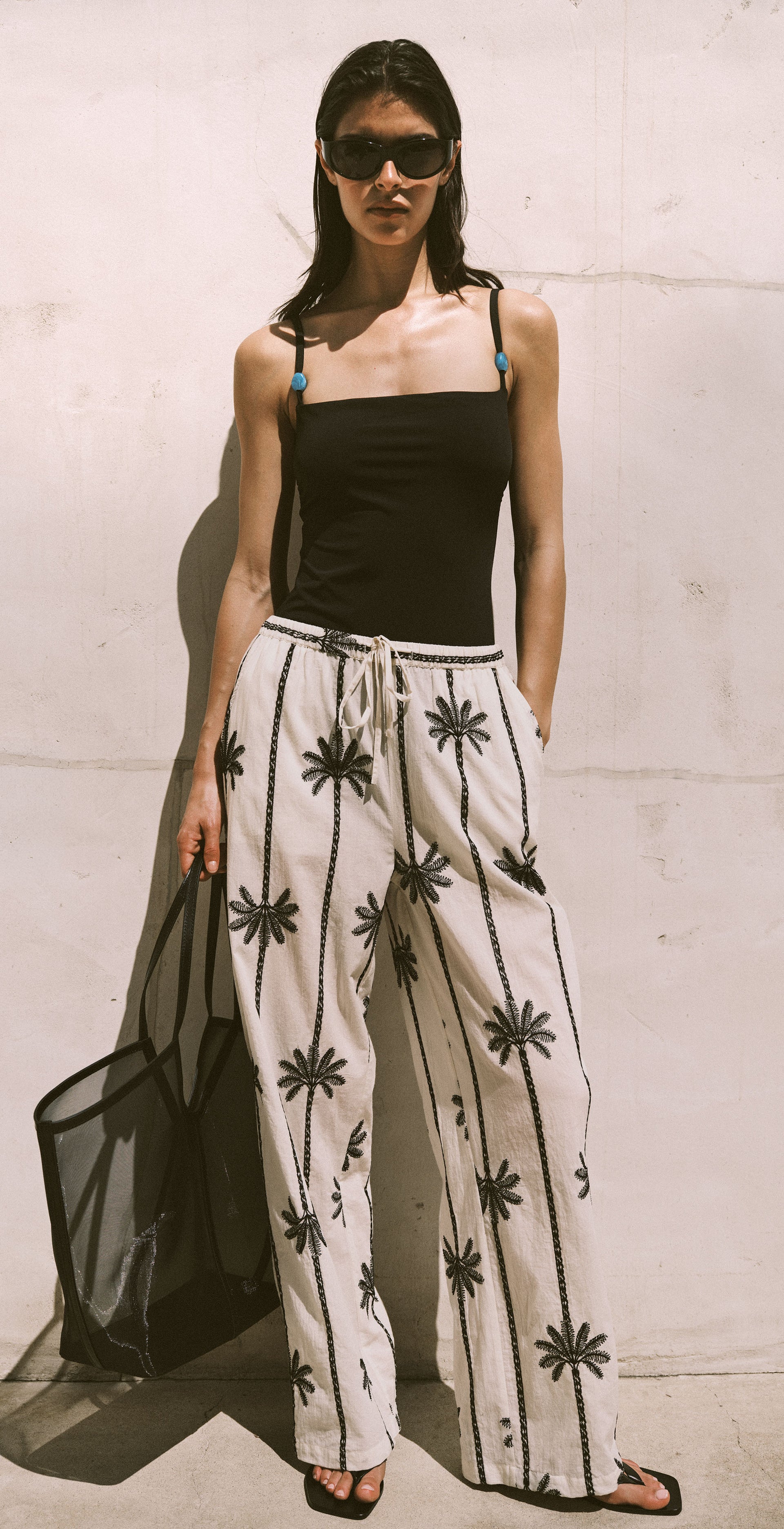 A three-panel lifestyle photo of a woman modeling a black camisole top and white wide-leg pants with a black palm tree print, posed against a light concrete wall with a large clay pot.