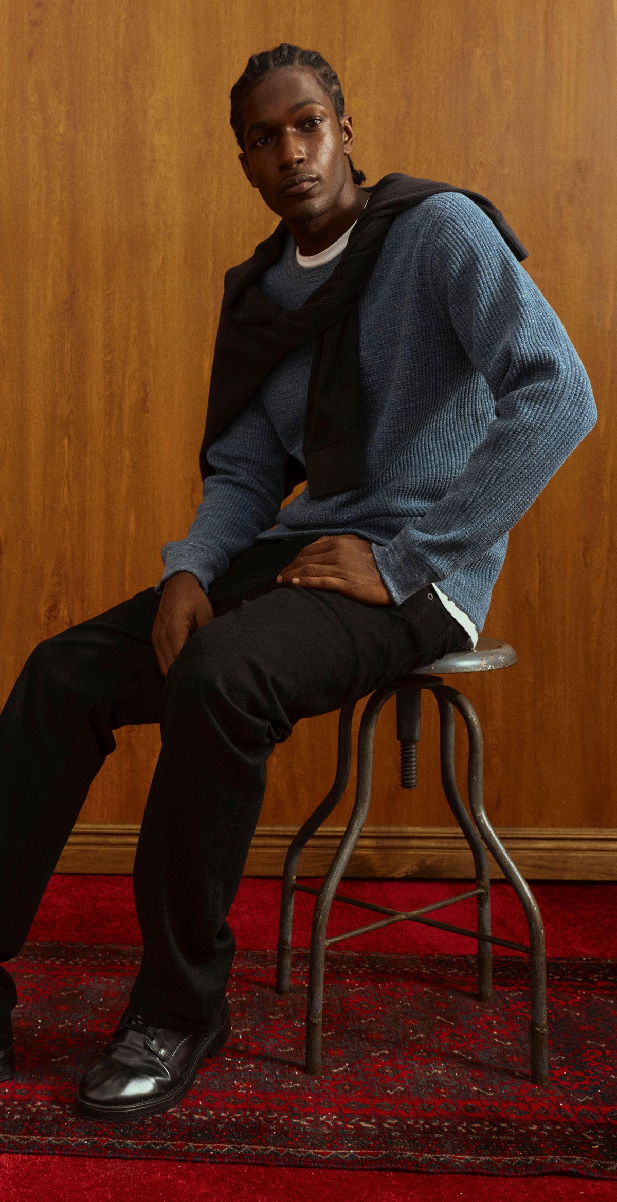 A man wearing a blue textured knit sweater, black hoodie, and black pants sits on a stool against a warm wood-paneled background.