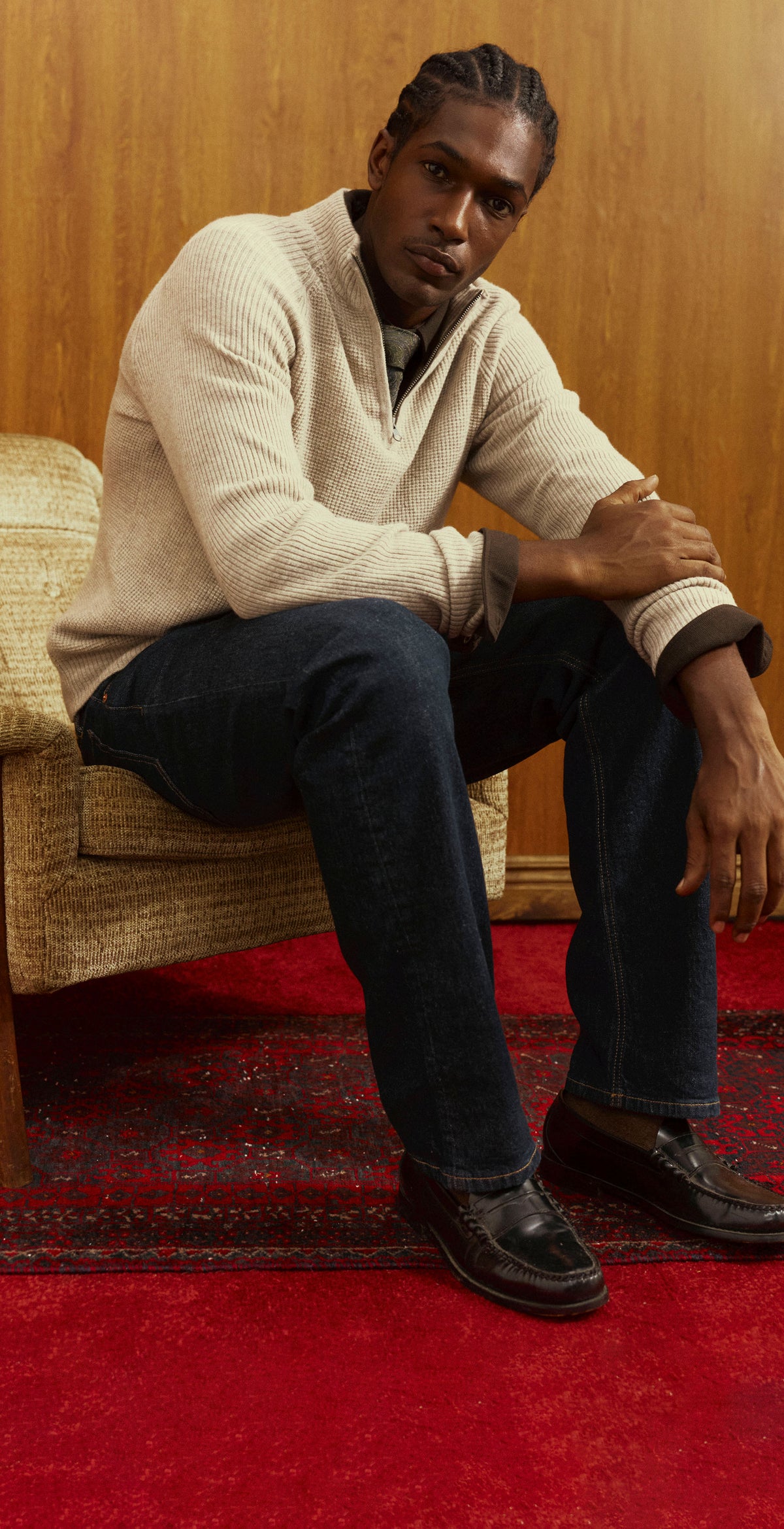 A split-screen lifestyle photo of a male model wearing a cream-colored quarter-zip sweater and dark blue jeans, posed against a wood-paneled wall and sitting in a woven armchair on a red patterned rug.