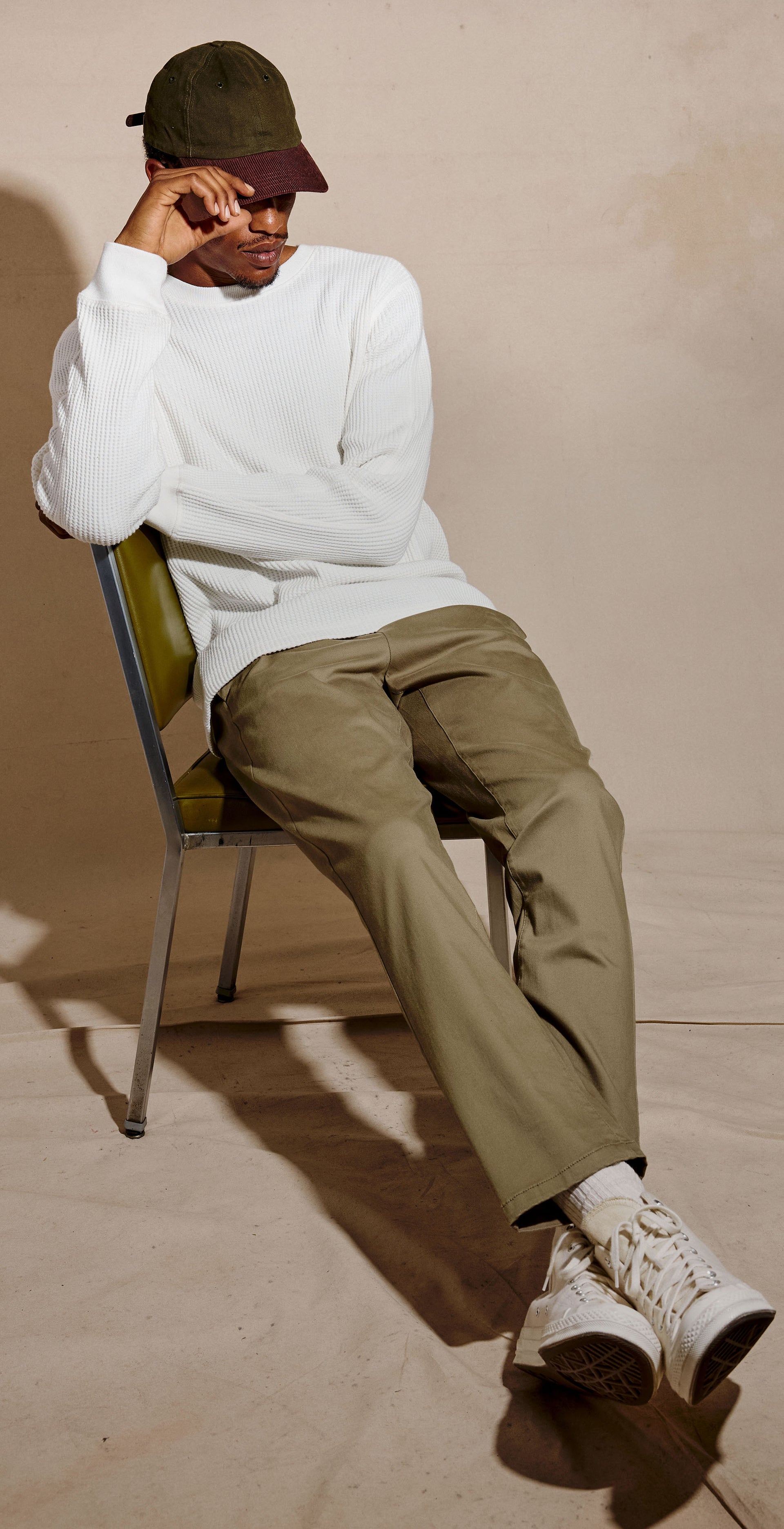 A three-panel fashion photo showing a man modeling a white long-sleeve crewneck shirt, olive green pants, white sneakers, and a dark green cap while sitting on and standing beside chairs against a warm beige studio backdrop.