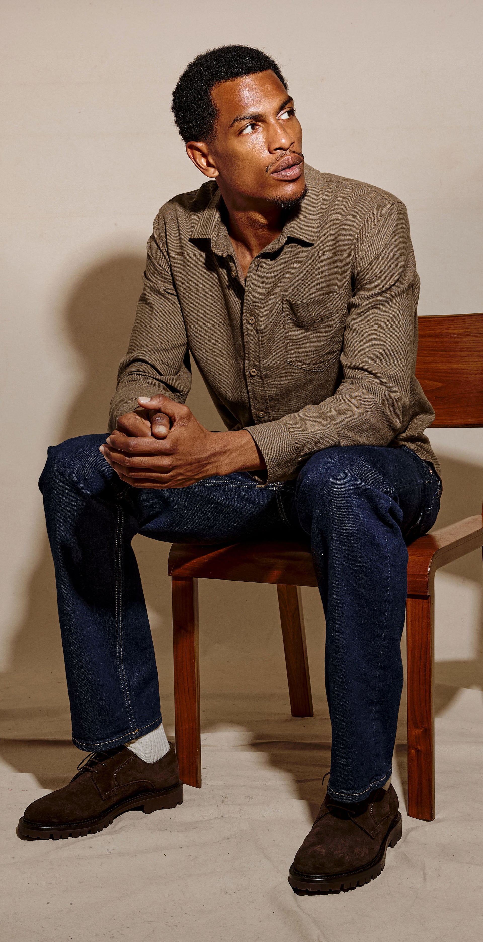 A man sits on a wooden chair wearing a brown button-up shirt, dark blue jeans, and brown shoes against a textured beige studio backdrop.