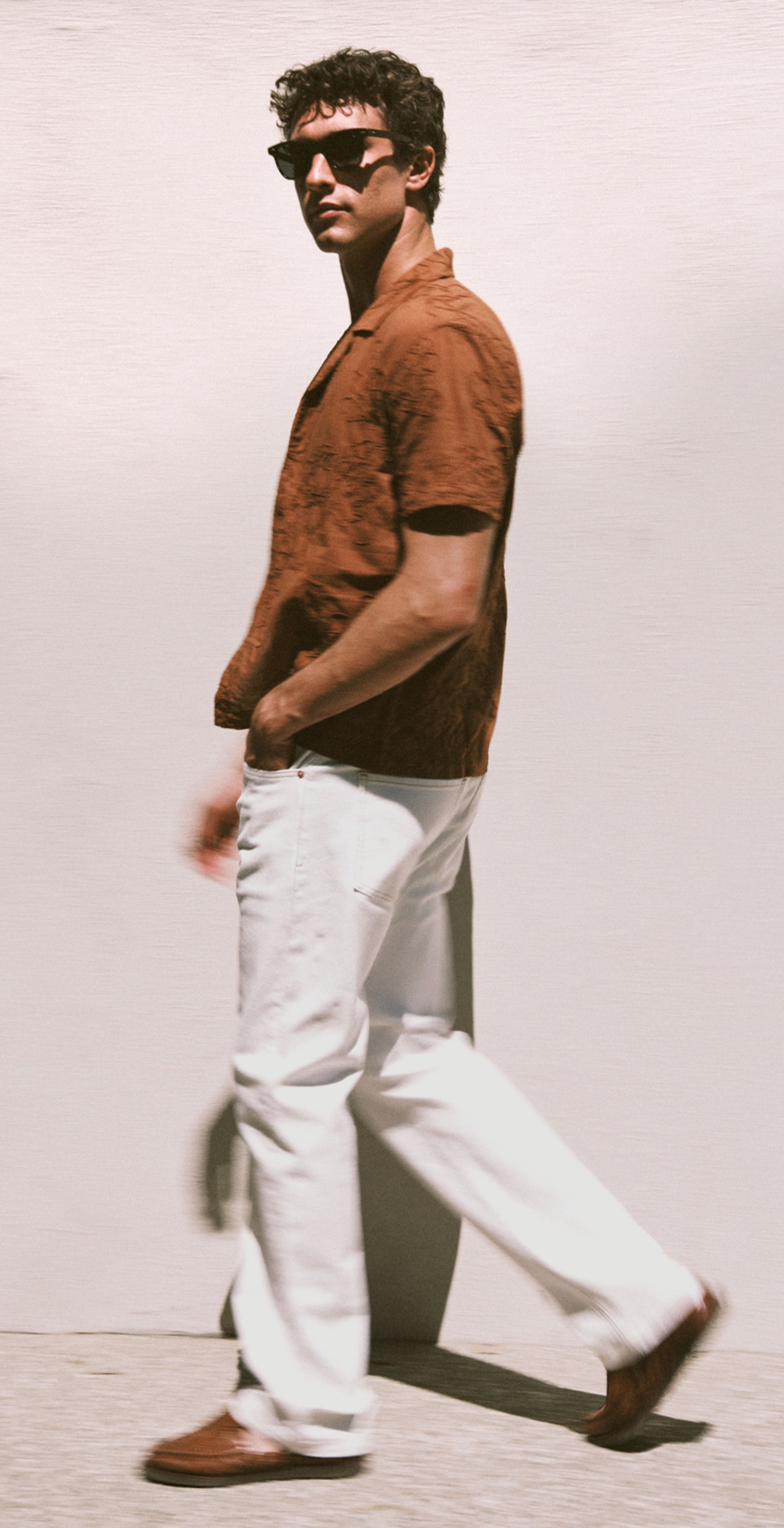 Man wearing sunglasses and a brown shirt standing against a wall.