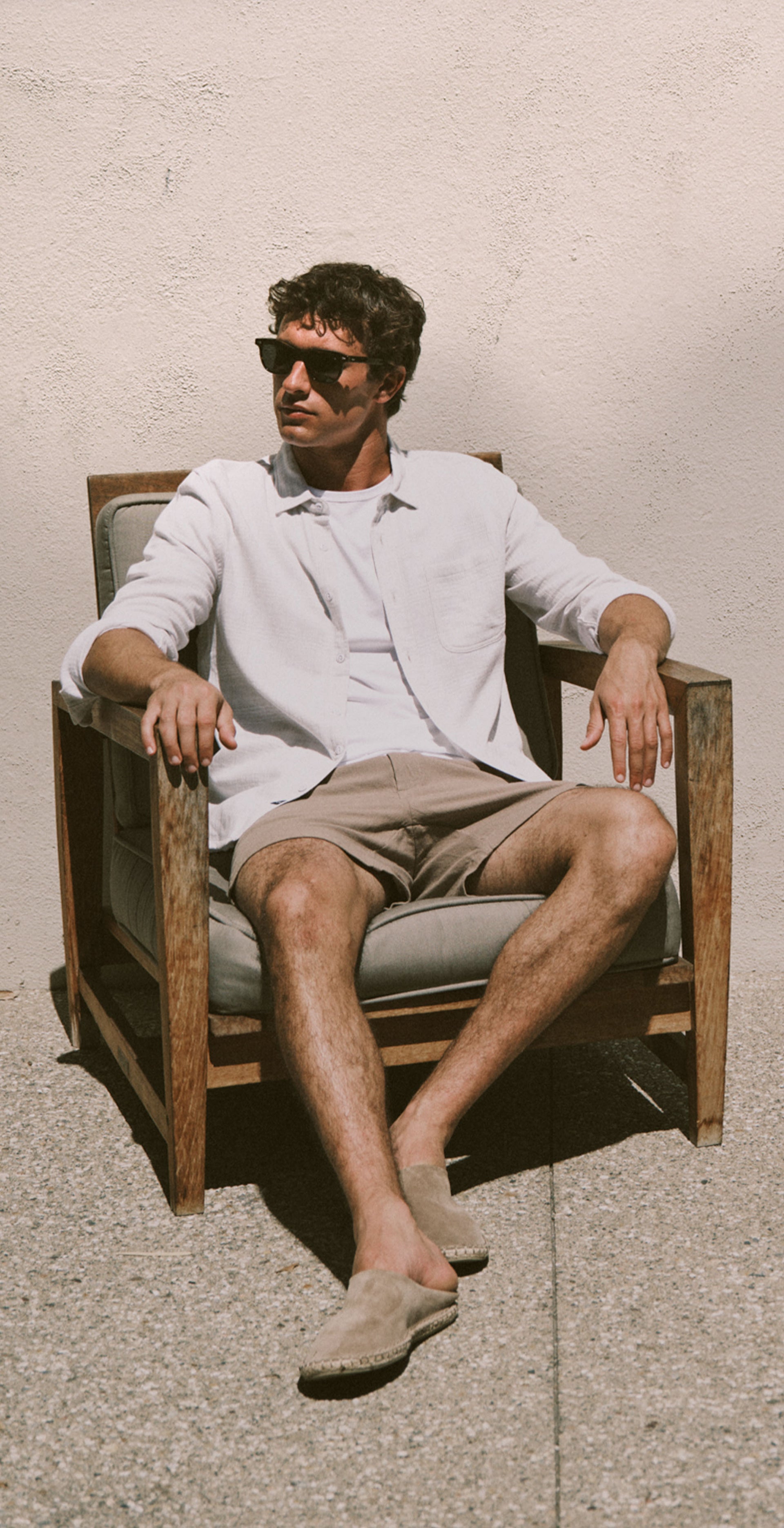 A three-panel lifestyle photo of a man wearing a light-colored linen button-up shirt, beige shorts, and sunglasses, shown standing, in a close-up, and sitting in a wooden chair against a textured beige wall.