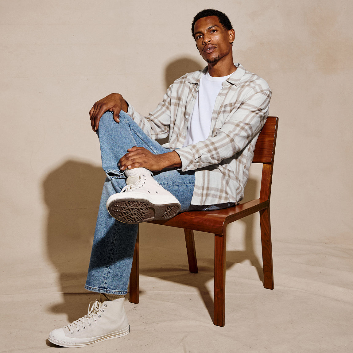 A man sits on a wooden chair wearing a beige and white plaid flannel shirt over a white t-shirt, blue jeans, and white high-top sneakers against a warm beige studio backdrop.