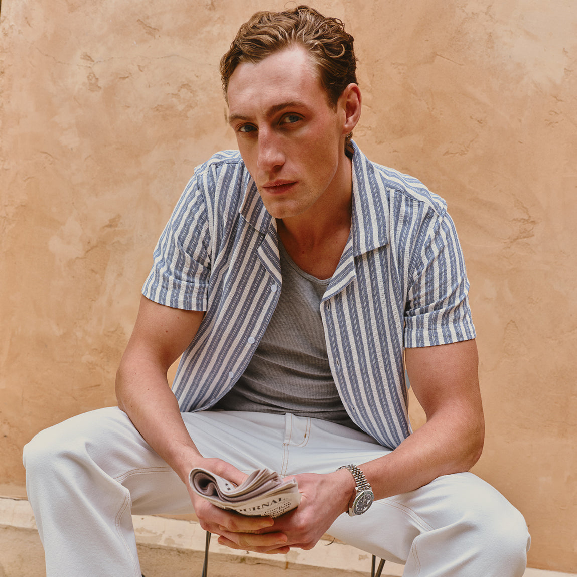 Man sitting against a beige wall wearing a striped shirt and holding a newspaper.