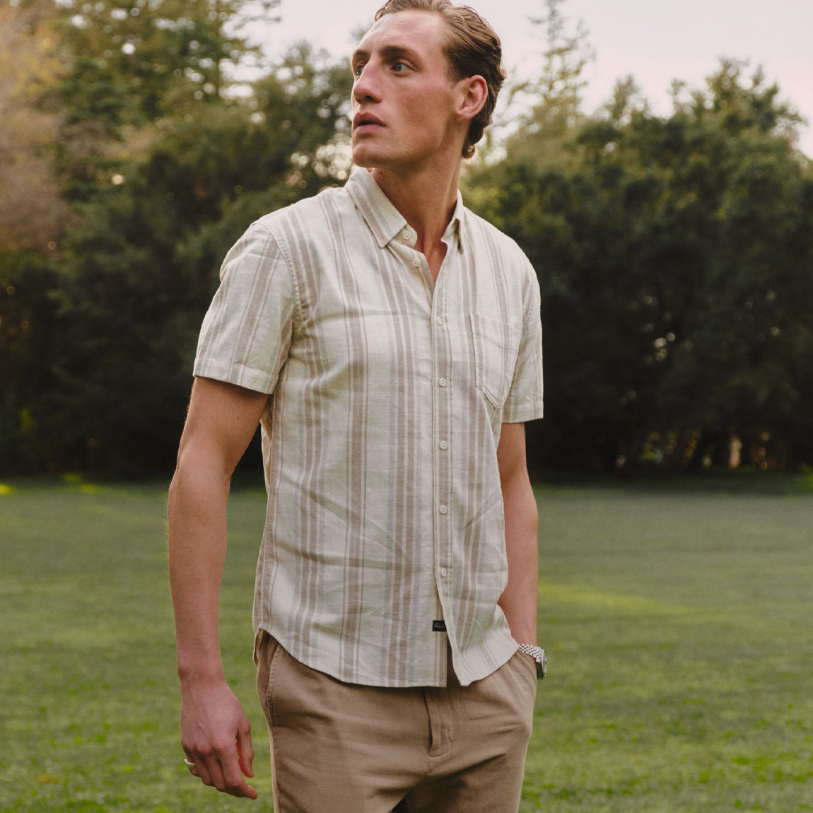 A man standing outdoors on a grassy lawn wearing a short-sleeve button-up shirt with vertical beige and white stripes, paired with khaki pants.