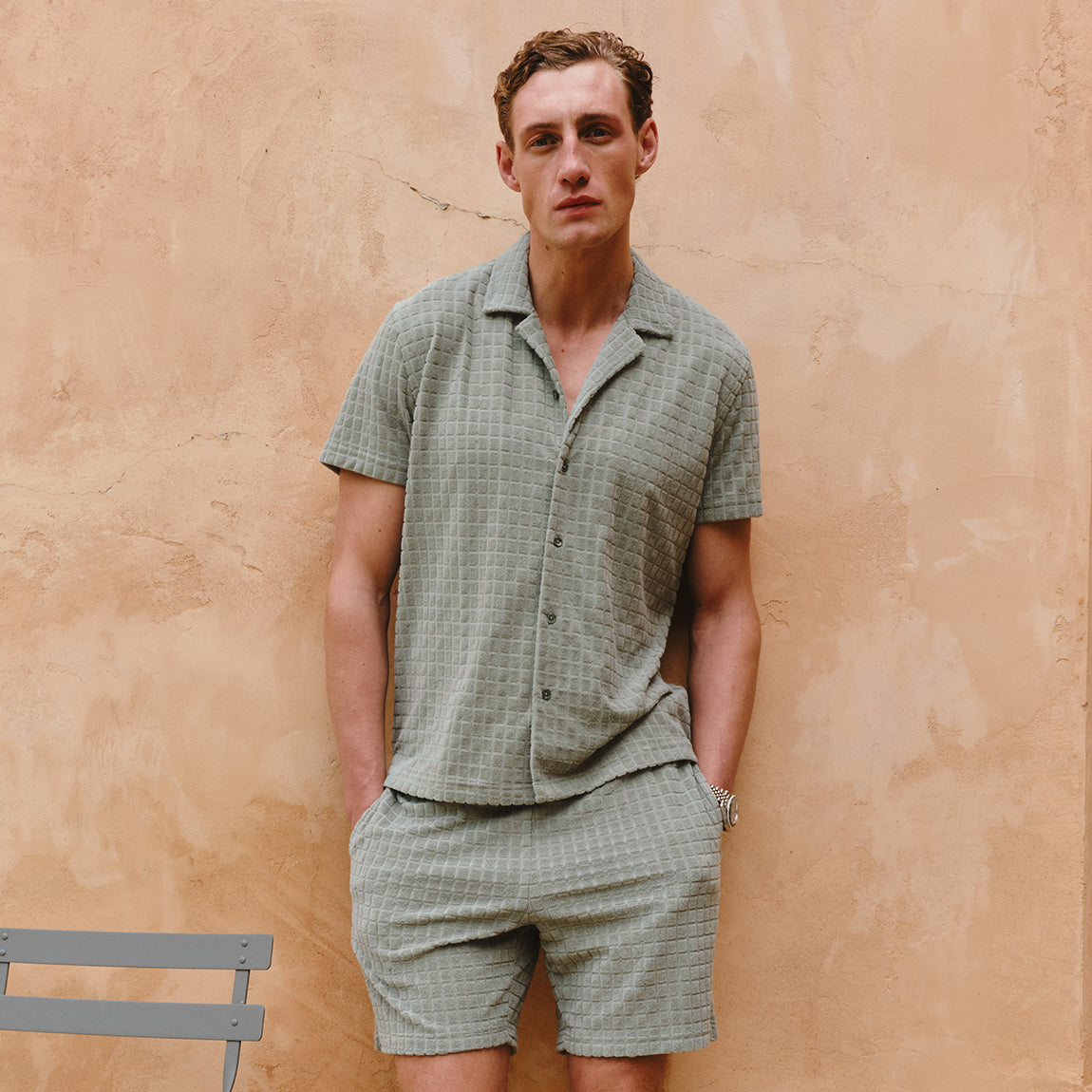 Man wearing a green checkered shirt and shorts against a beige wall.