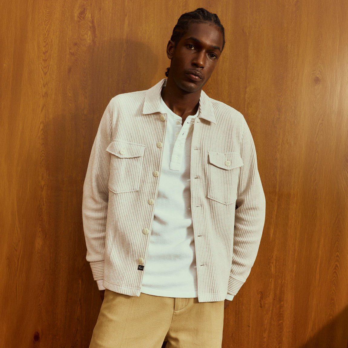 A man wearing a cream ribbed button-up overshirt over a white henley shirt and beige trousers, standing against a warm wood-paneled wall.