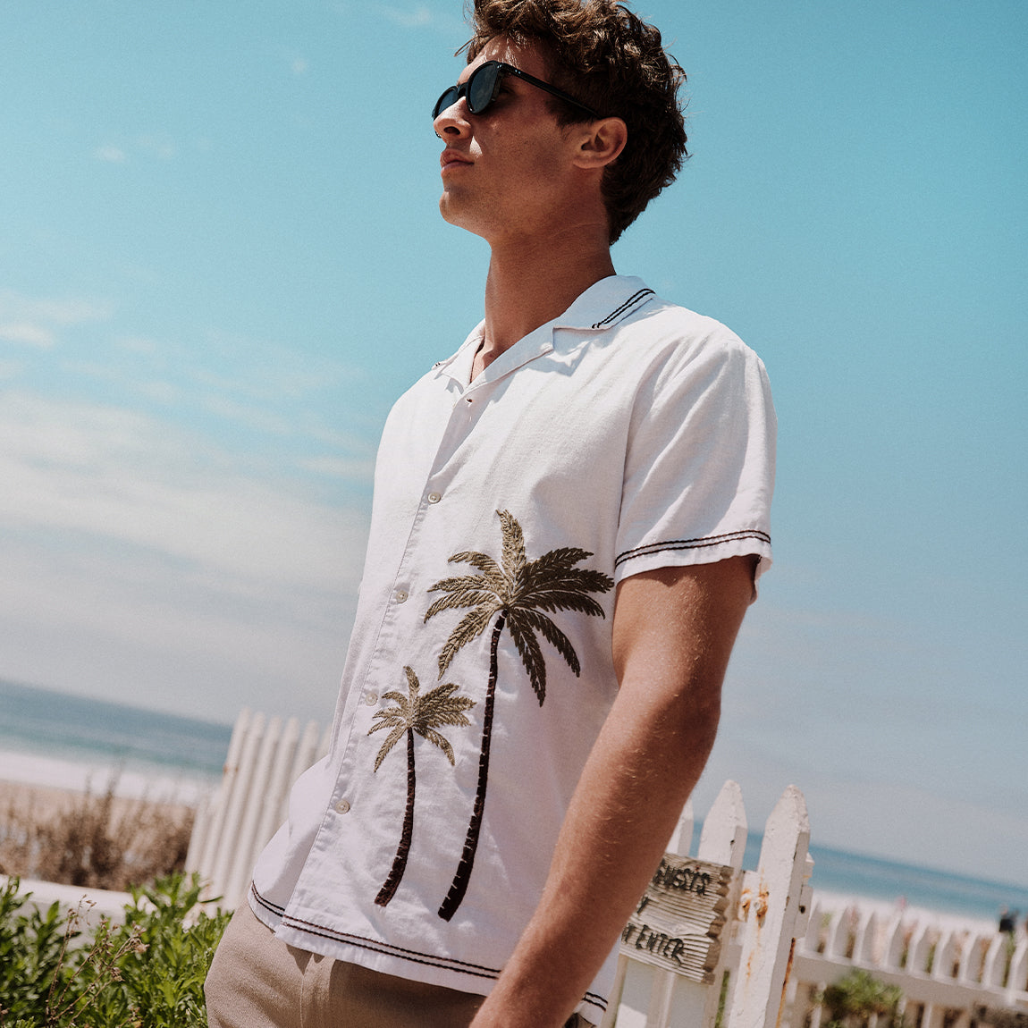 Man wearing a white polo shirt with palm tree design on a beach.