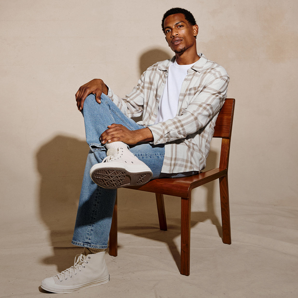 A man sits on a wooden chair wearing a beige and white plaid button-up shirt over a white t-shirt, blue jeans, and white high-top sneakers against a textured beige backdrop.