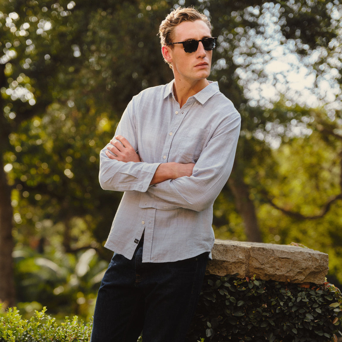 A man wearing a light gray long-sleeve linen button-up shirt and dark sunglasses, standing outdoors with arms crossed in front of a stone wall and green trees.
