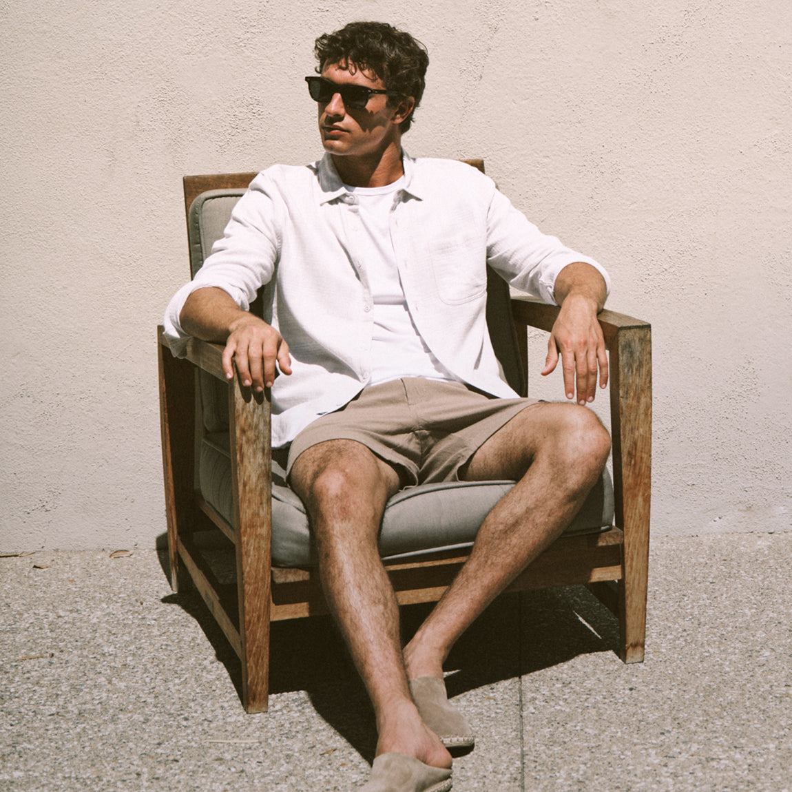 Man sitting on a chair wearing sunglasses and casual clothing against a plain wall.
