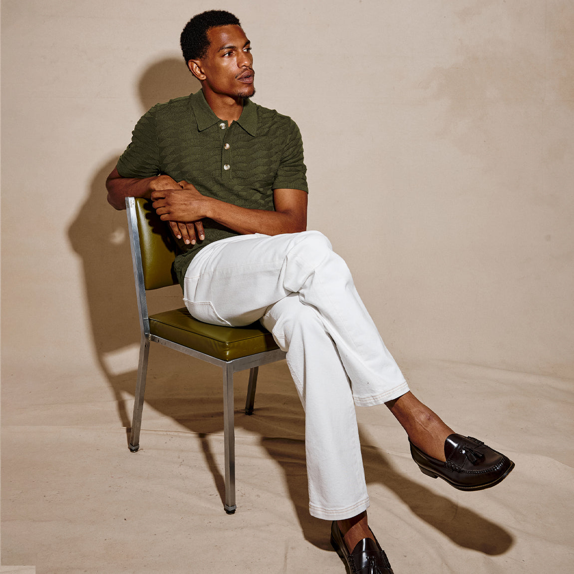 A man sits on a green and chrome chair wearing a dark olive green striped polo shirt, white trousers, and black loafers against a beige studio backdrop.