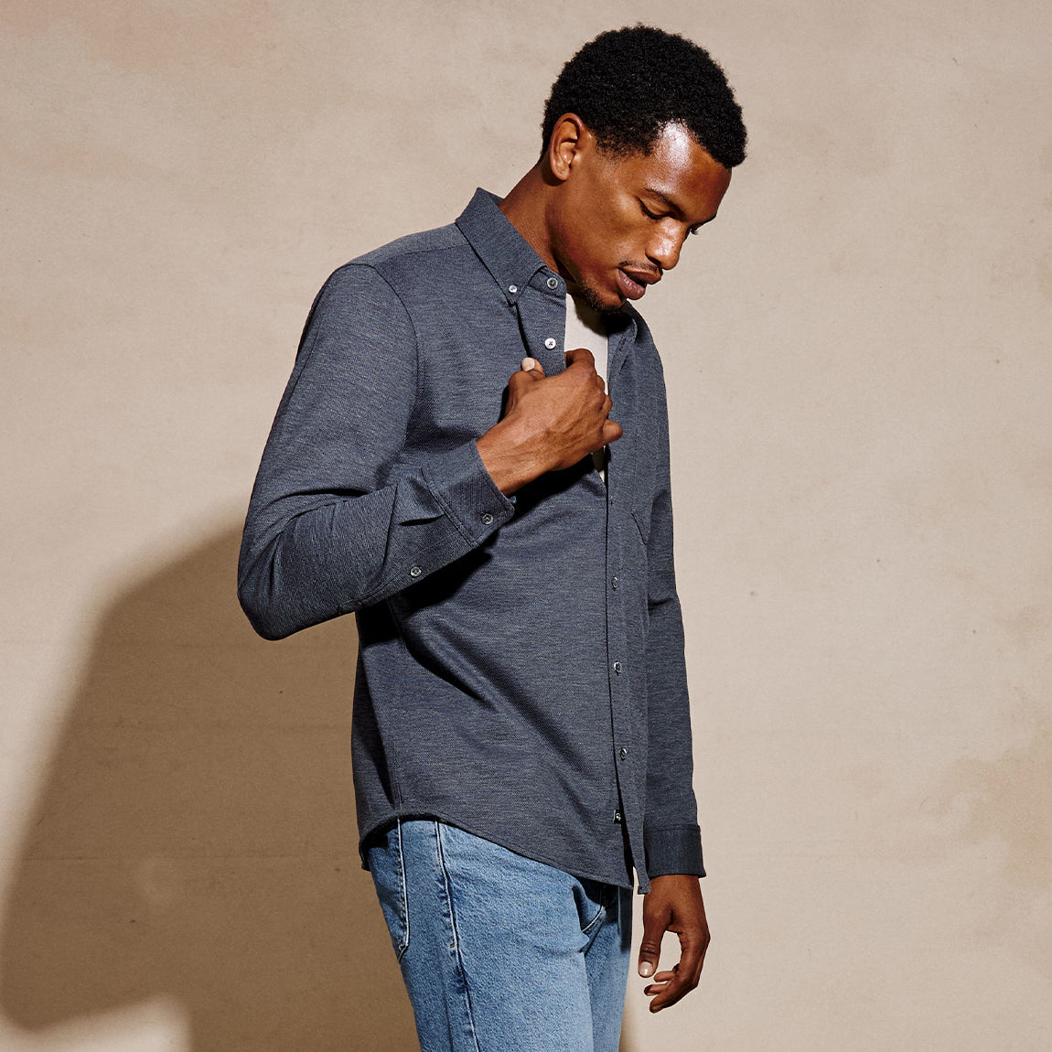 A man wearing a dark gray heathered long-sleeve button-down shirt and blue jeans, standing against a textured beige wall.