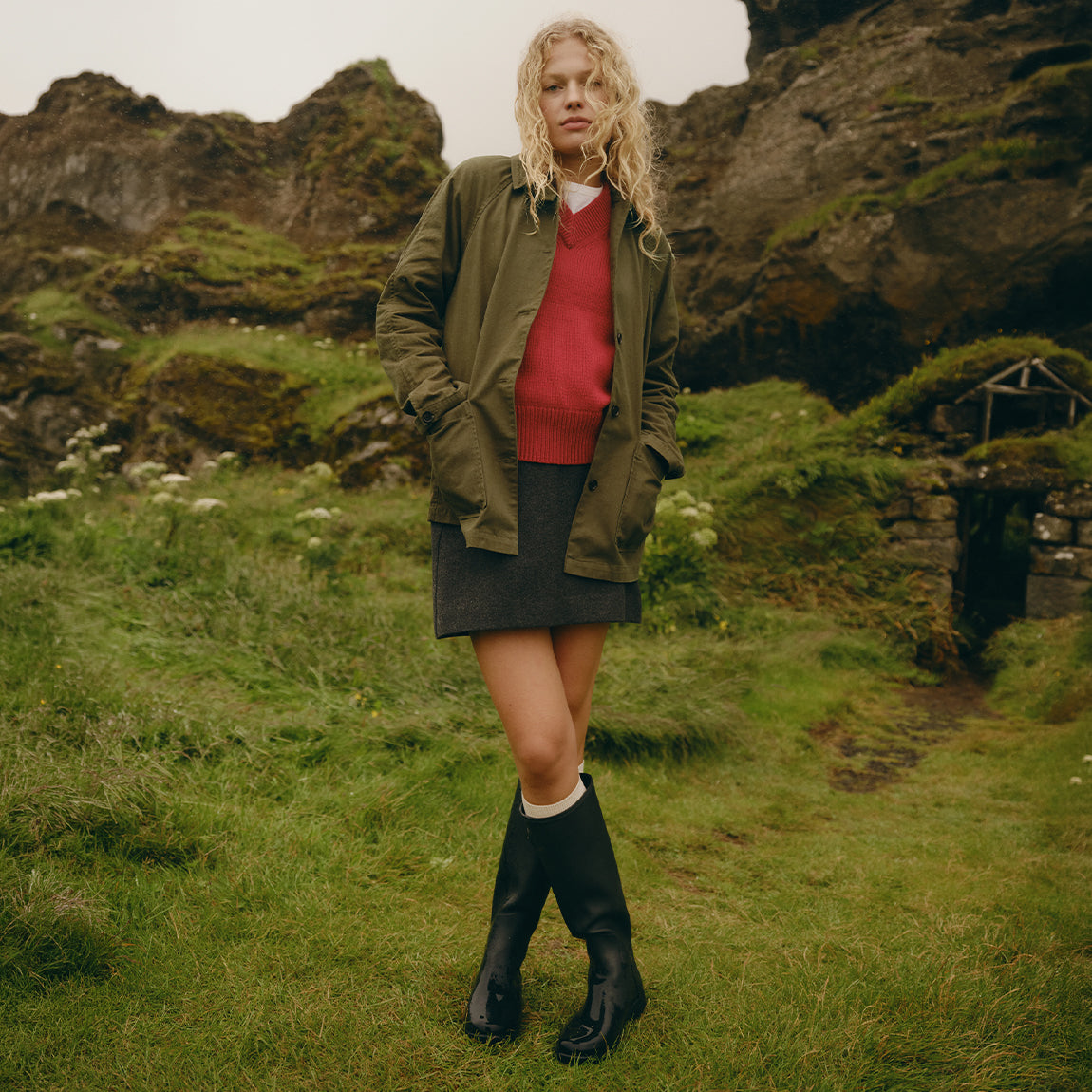 A woman stands outdoors in a grassy, rocky landscape wearing an olive green jacket over a red sweater and dark skirt, with black rain boots.
