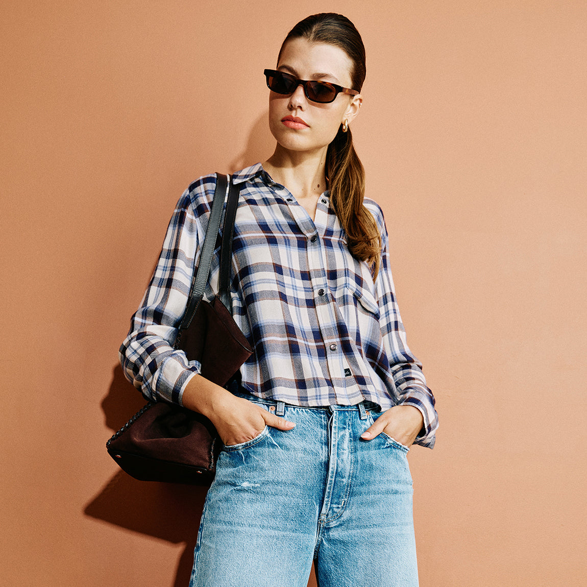 A woman wearing a blue, white, and navy plaid button-up shirt, light-wash blue jeans, and dark sunglasses, carrying a dark brown leather shoulder bag against a solid terracotta background.