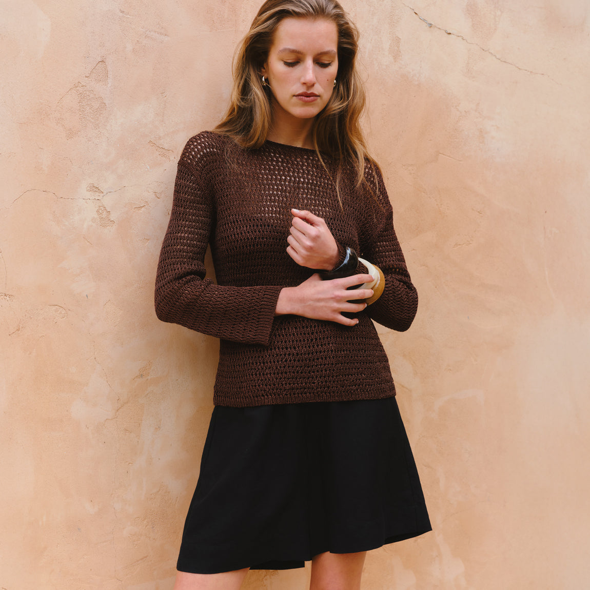 Woman wearing a brown knitted sweater and black skirt against a beige wall.