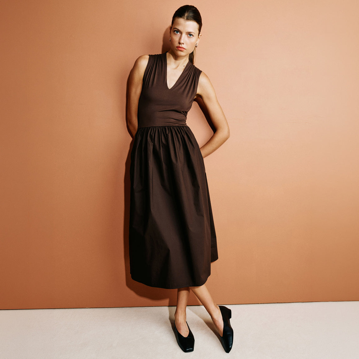A woman modeling a sleeveless dark brown midi dress with a V-neckline and gathered waist, standing against a solid peach-colored studio backdrop.