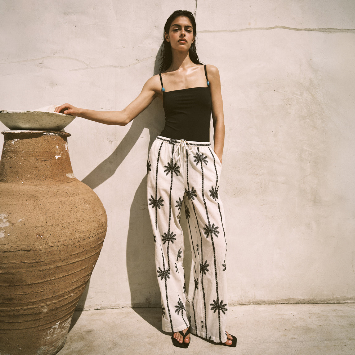 Woman wearing a black top and white pants with black patterns leaning against a large pot.