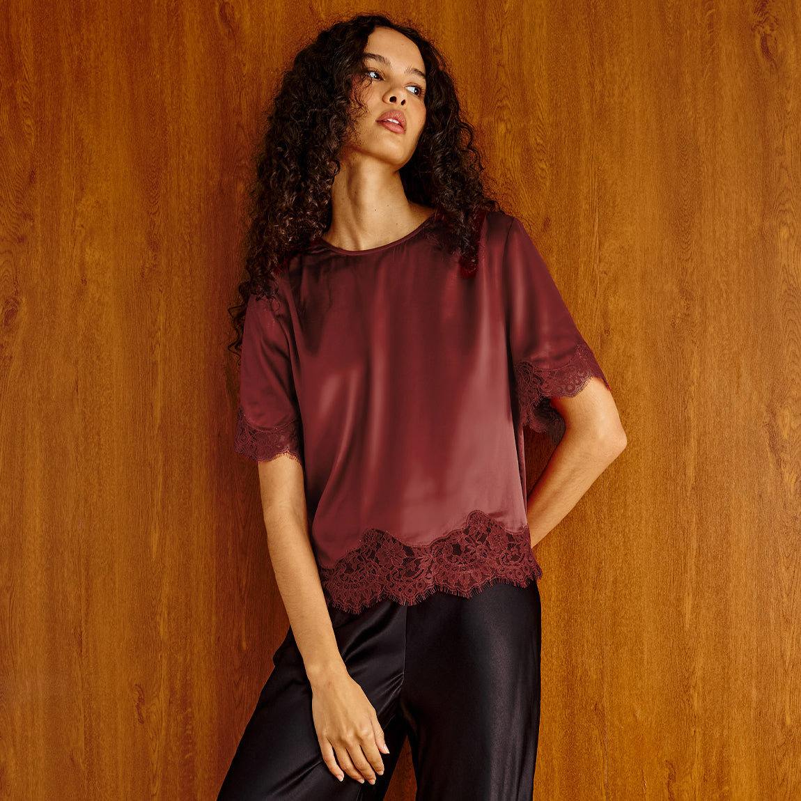 A woman with curly dark hair models a short-sleeve burgundy satin top with lace trim at the hem and cuffs, paired with black satin pants, set against a warm wood-paneled background.