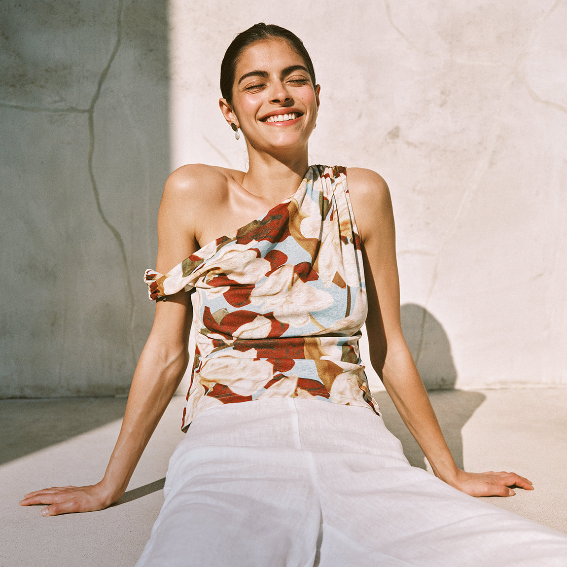 Woman wearing a floral top and white pants sitting against a light-colored wall.