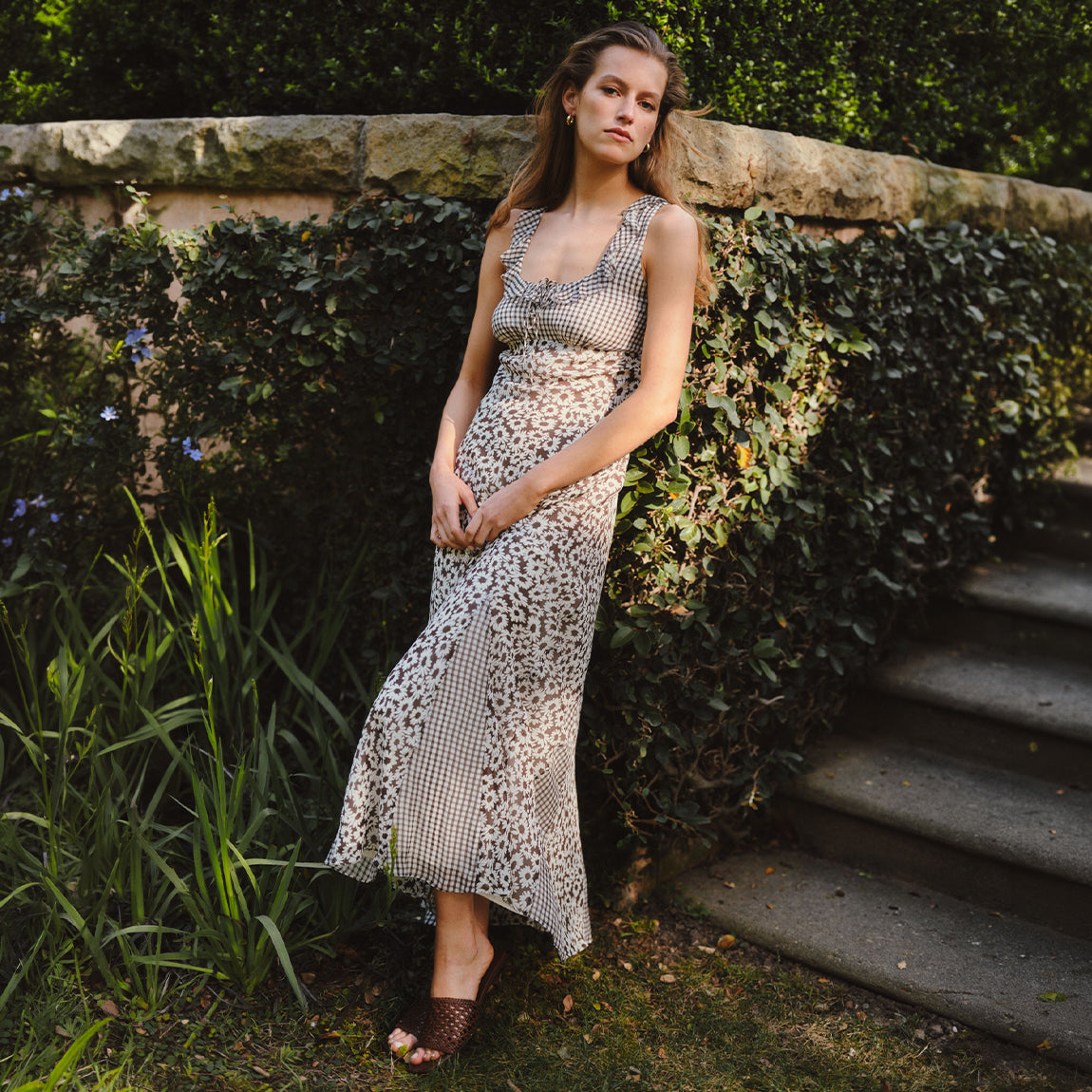 A woman wearing a sleeveless leopard-print maxi dress and brown woven sandals, posing outdoors against a stone wall with green ivy and garden plants.