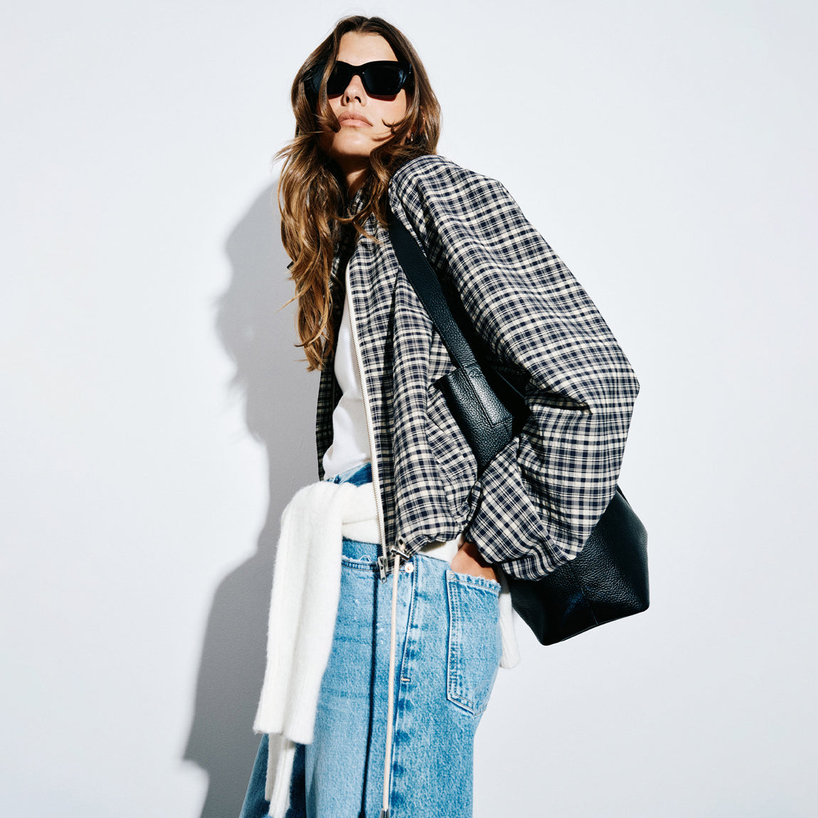 A woman wearing a black and white checkered bomber jacket, blue jeans, a white shirt, and black sunglasses, carrying a black leather shoulder bag against a white background.