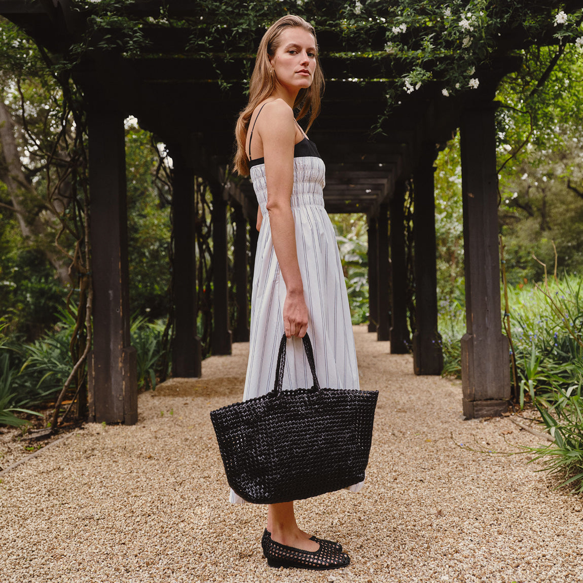 A woman stands on a gravel path under a wooden pergola, wearing a white sleeveless midi dress with a black bodice and holding a large black woven tote bag.