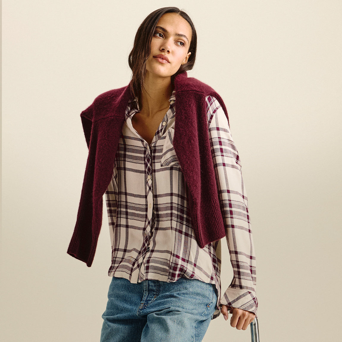 A woman wearing a burgundy knit sleeveless vest over a beige and purple plaid button-down shirt with blue jeans, photographed against a plain beige studio background.