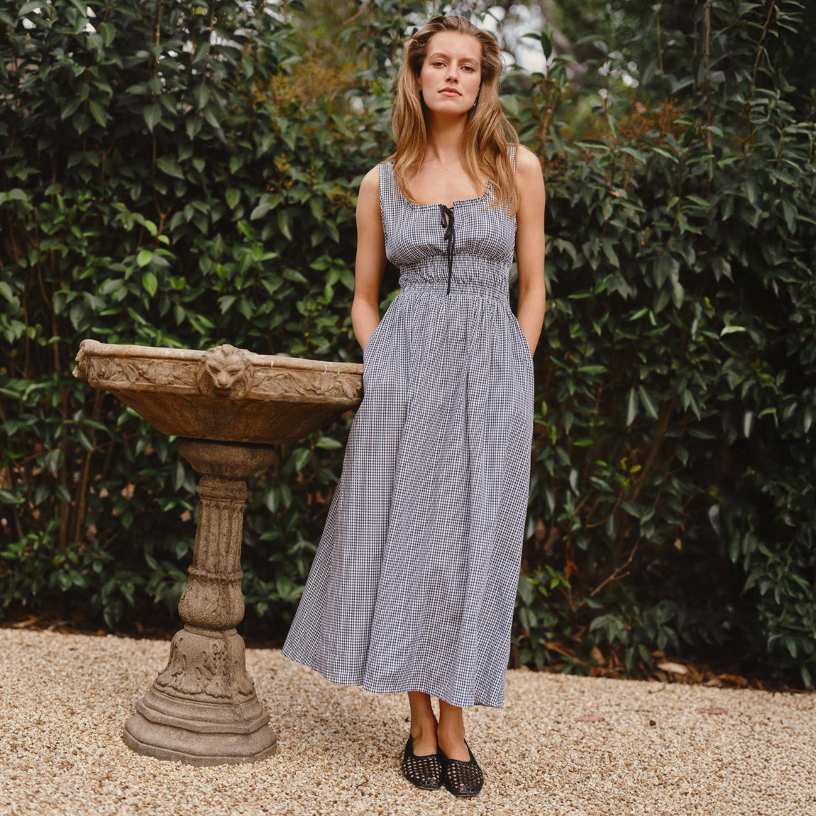 Woman in a blue checkered dress standing next to a stone fountain in a garden.