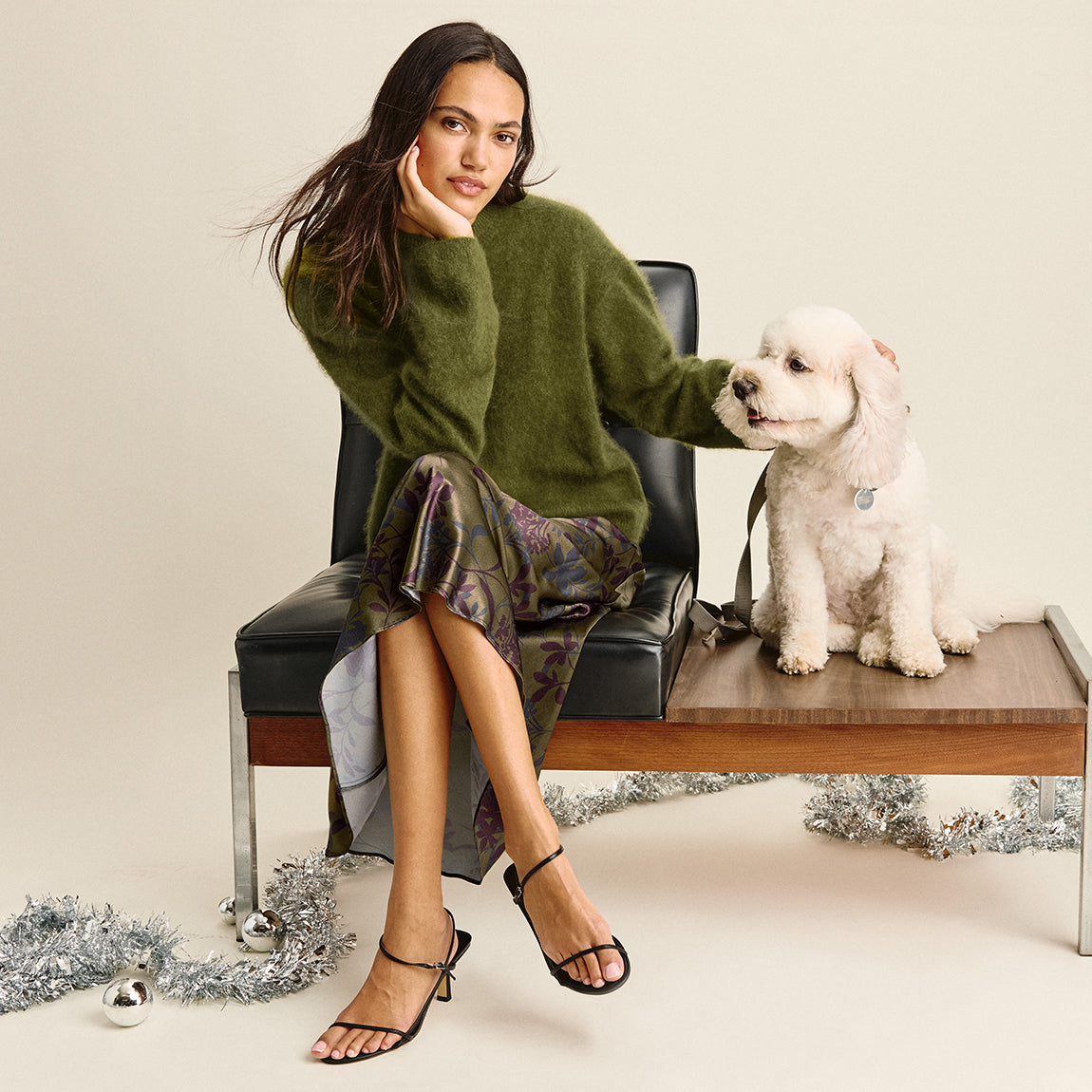 A woman sits on a black chair next to a small white dog on a wooden bench, wearing an oversized olive green knit sweater, a patterned skirt, and black strappy heels, against a warm off-white background with silver tinsel on the floor.
