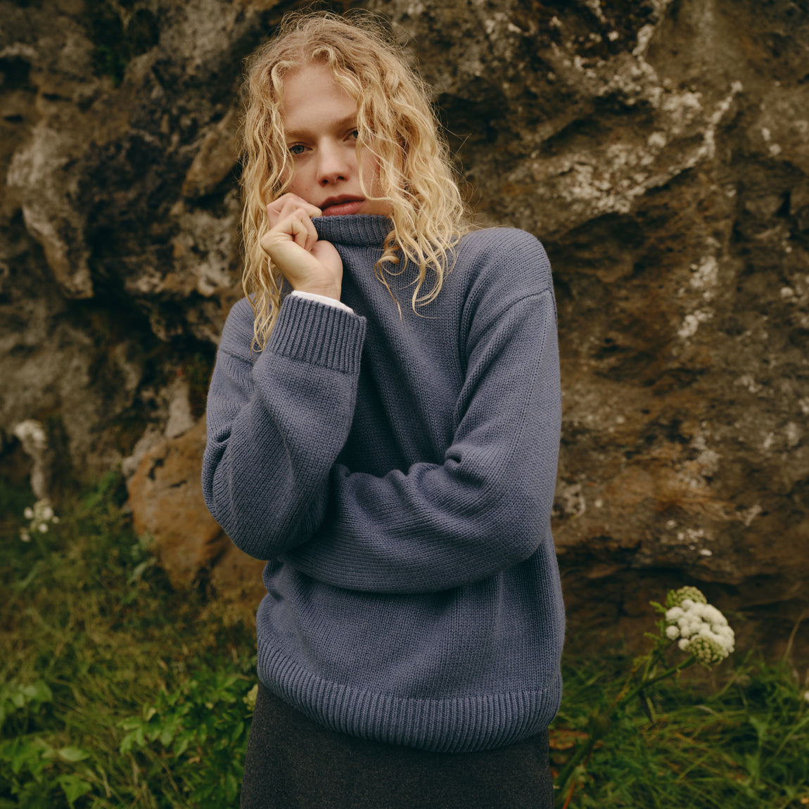 A woman with curly blonde hair wearing a slate blue turtleneck sweater, posing in front of a large rock wall outdoors.