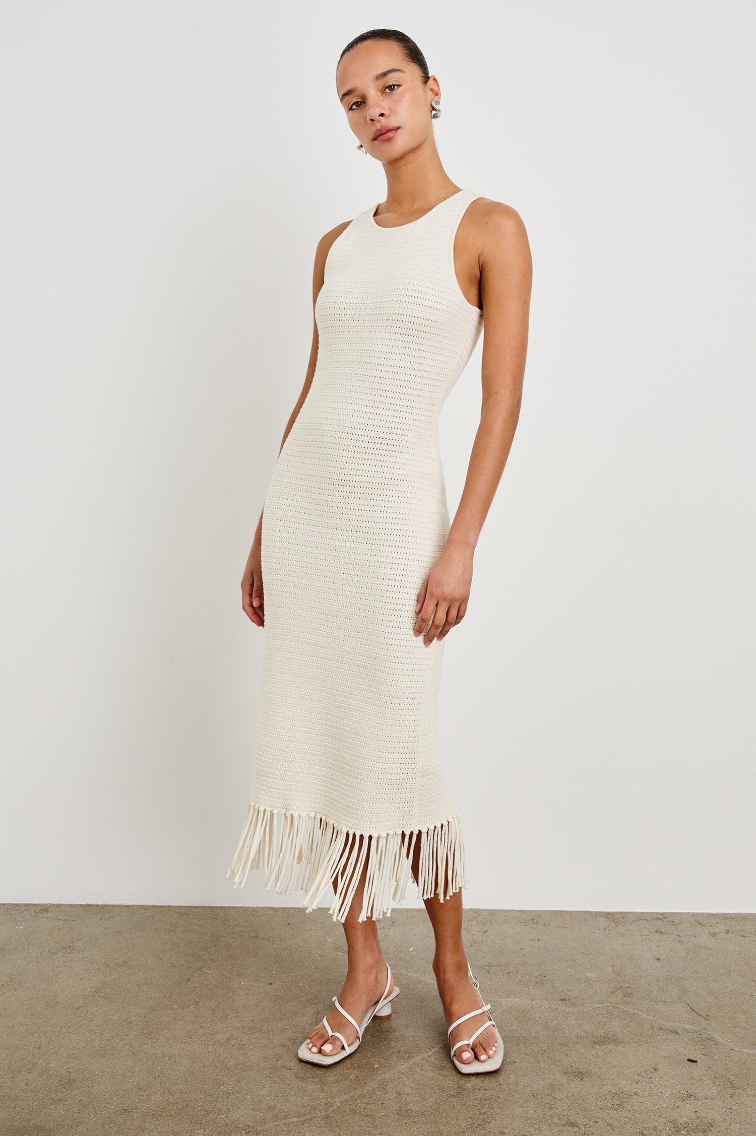 Front view of female model wearing our sleeveless, Nia Midi Dress in color Ecru, featuring crochet structure and fringe details at the hem.